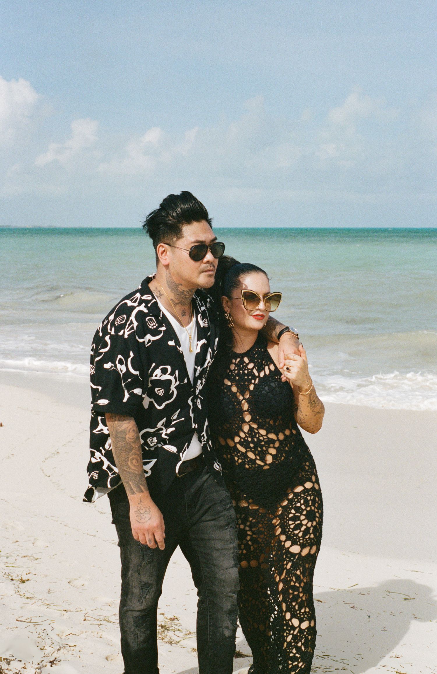 Fashionable couple posing on a tropical beach, man in floral shirt, woman in black crochet dress.