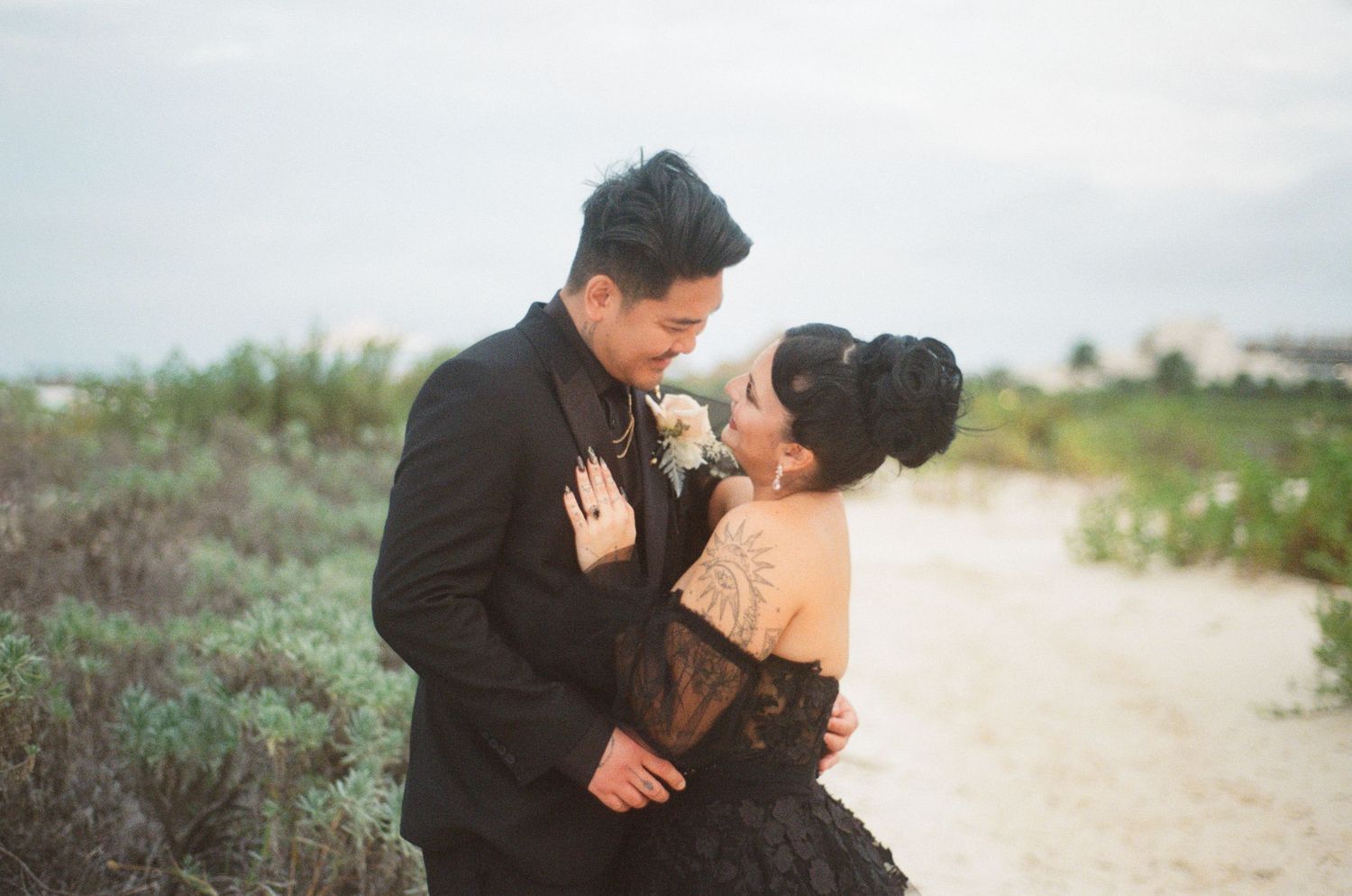 Bride and groom sharing intimate moment, black lace wedding gown with tattoo detail, beach dunes setting.