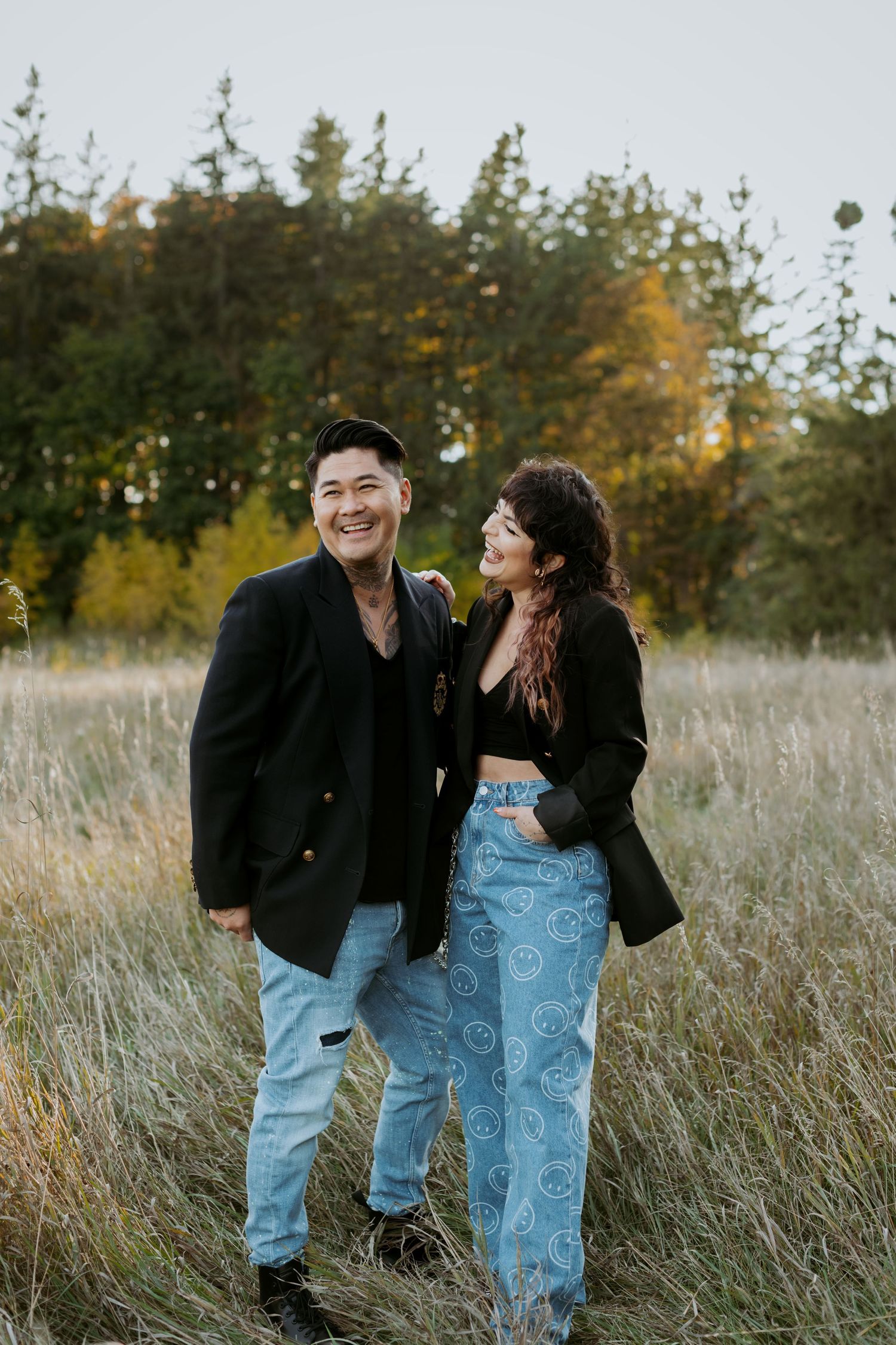 A joyful couple in matching black blazers and jeans laughing together in an autumn field.