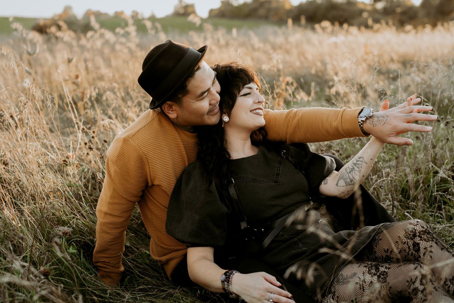 Couple sitting in golden field, woman laughing with arms outstretched, man in black hat embracing her at sunset.