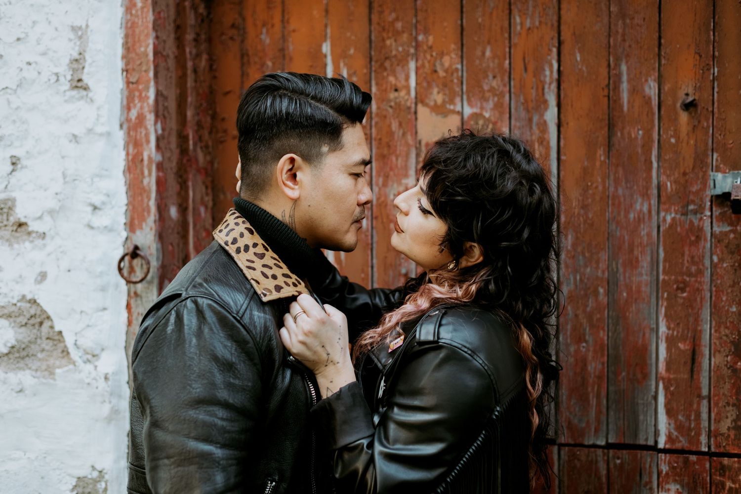 Stylish couple in black leather jackets facing each other by a vintage wooden door.