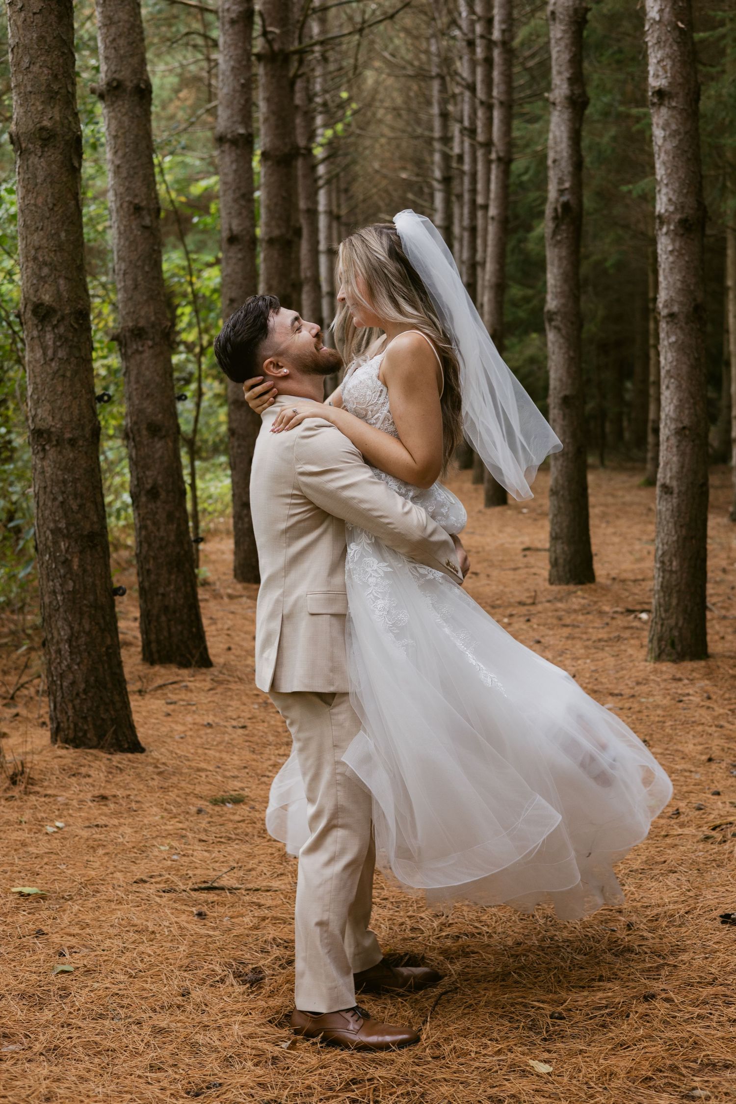 Groom lifts bride in a white lace gown and veil, sharing a kiss in a sunlit pine forest pathway.