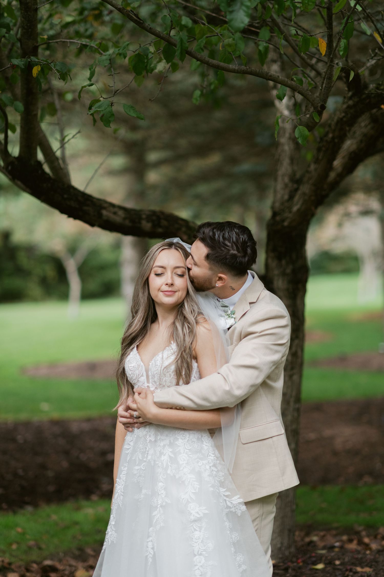 Groom in tan suit embracing bride in white lace gown under a tree in a green garden.