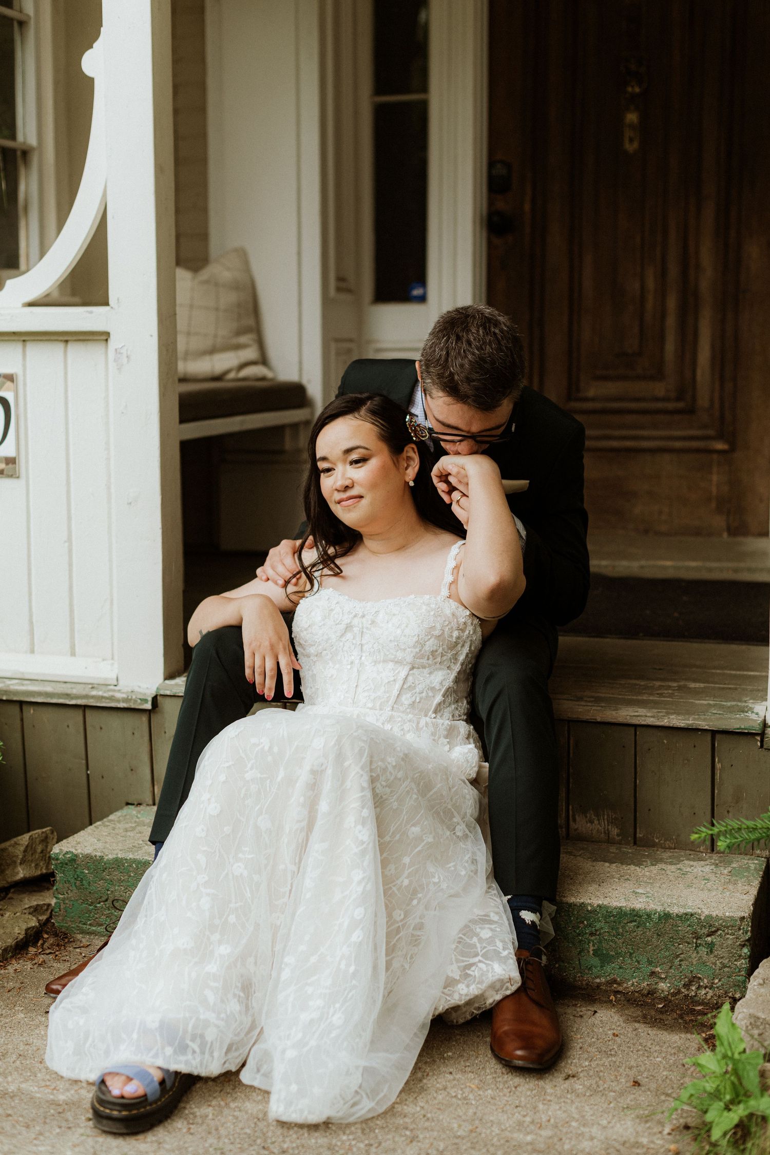 Bride in lace gown sits on porch steps as groom whispers in her ear tenderly.