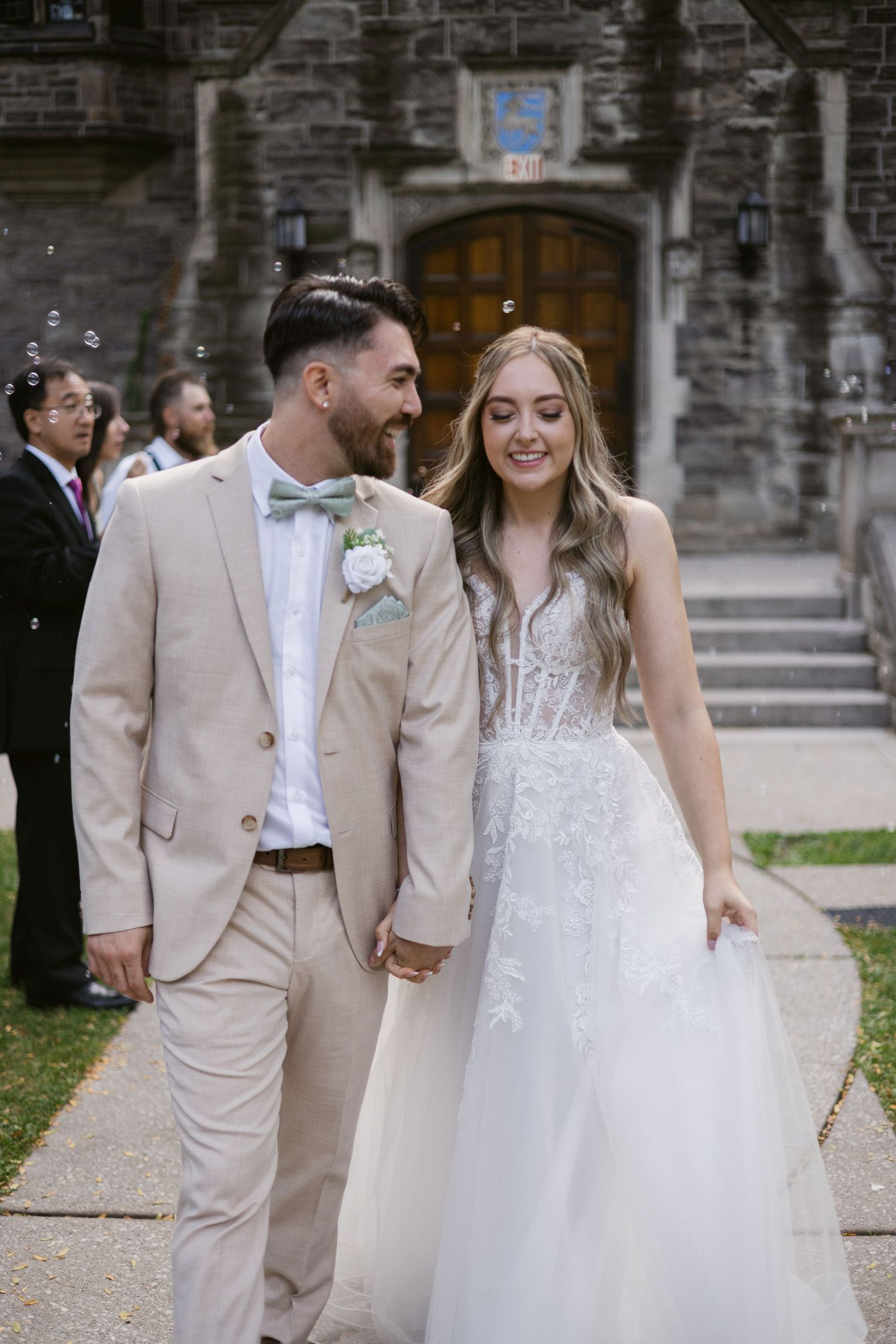 Groom in tan suit and bride in lace gown stroll hand-in-hand outside stone chapel.