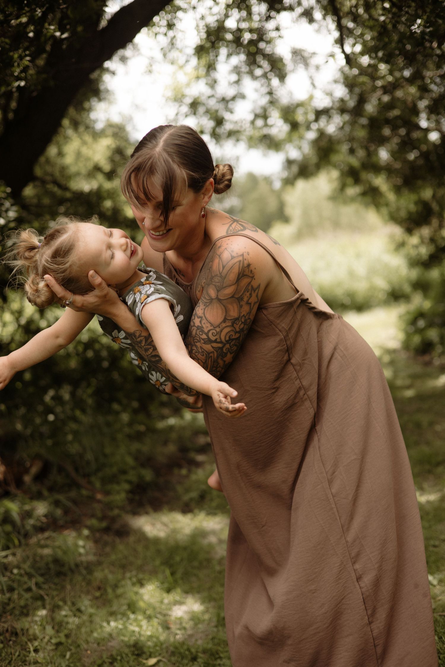 A tattooed mother in a brown dress playfully dips her laughing toddler outdoors.