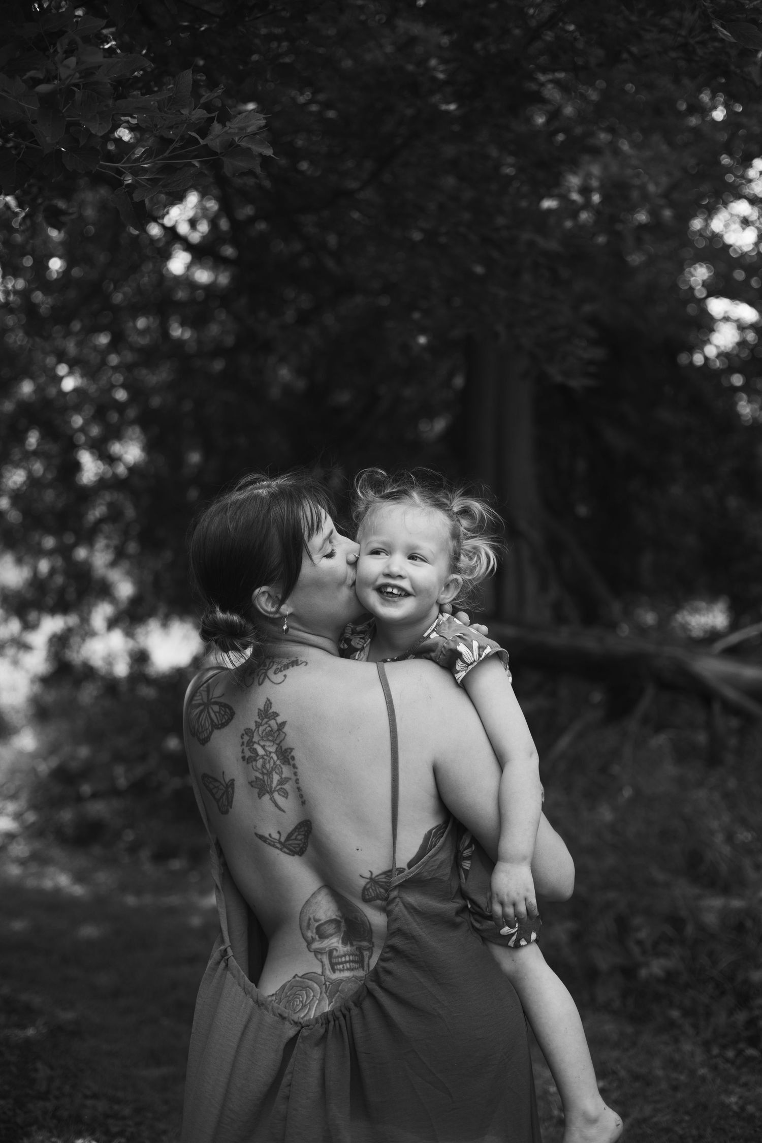 Black and white photo of a tattooed mother kissing her giggling toddler daughter in a forest.