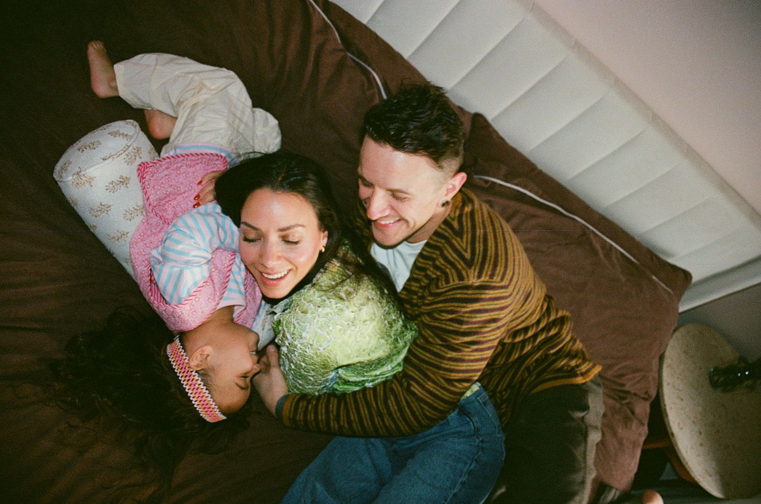 A couple smiles warmly while cuddling with their child and a cabbage on a brown sofa, viewed from above.