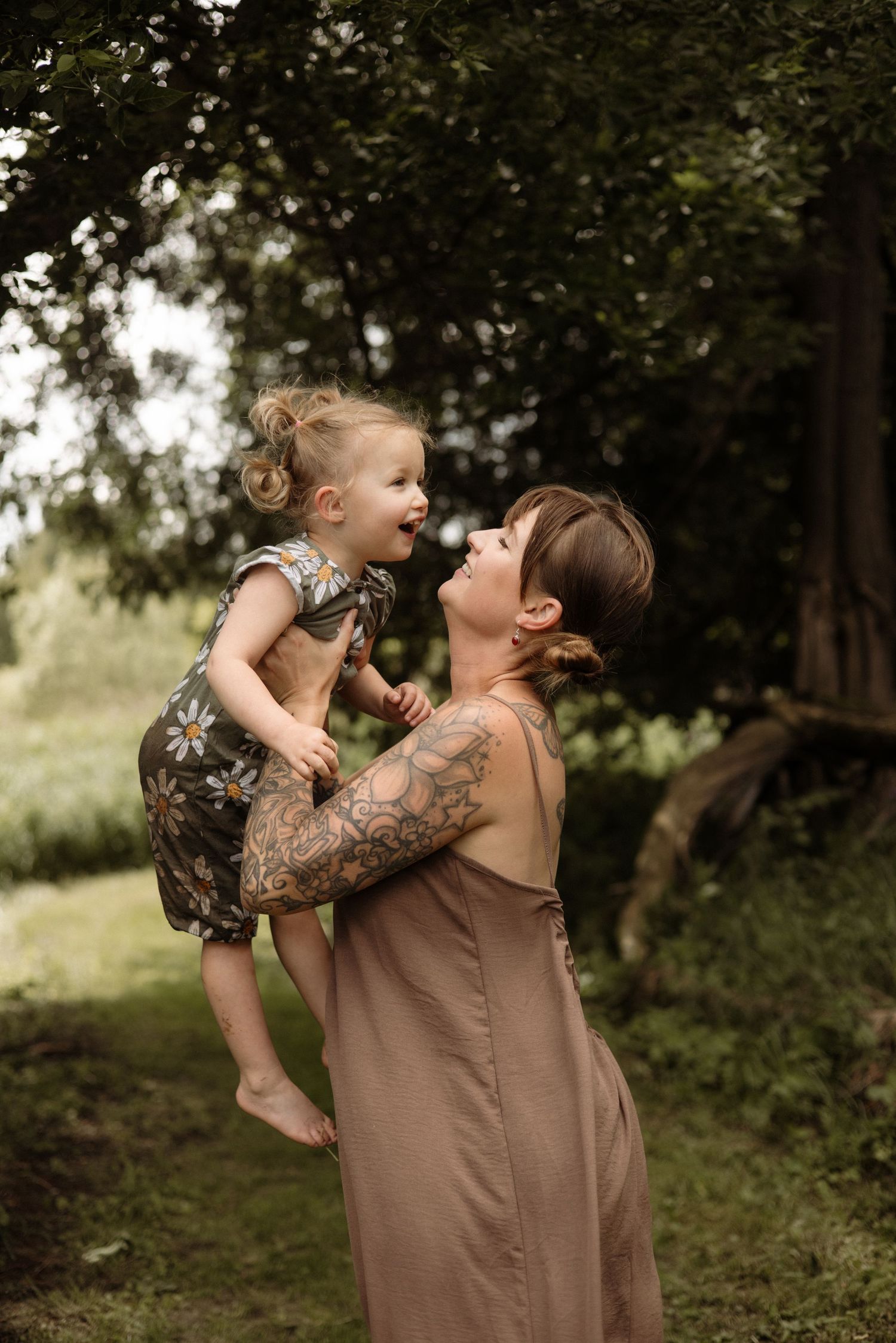 A tattooed mother lifts her giggling toddler face-to-face in a green garden.