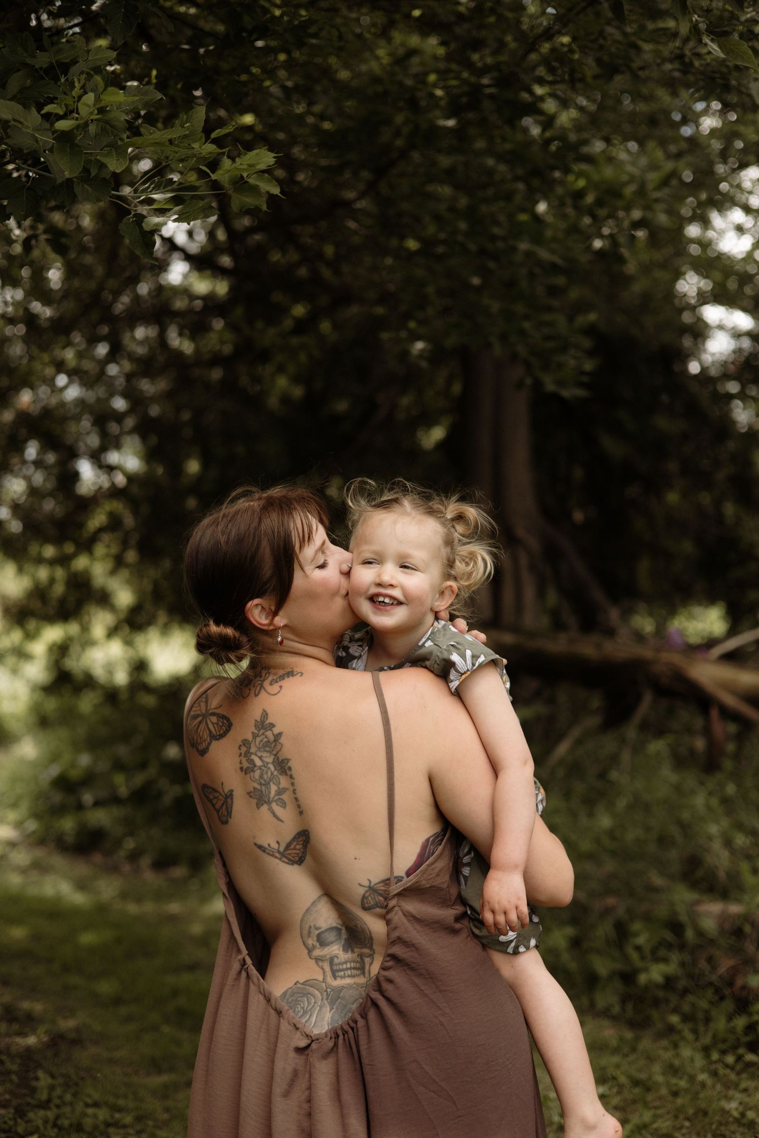 A tattooed mother in a backless brown dress kisses her smiling toddler daughter in a green forest.