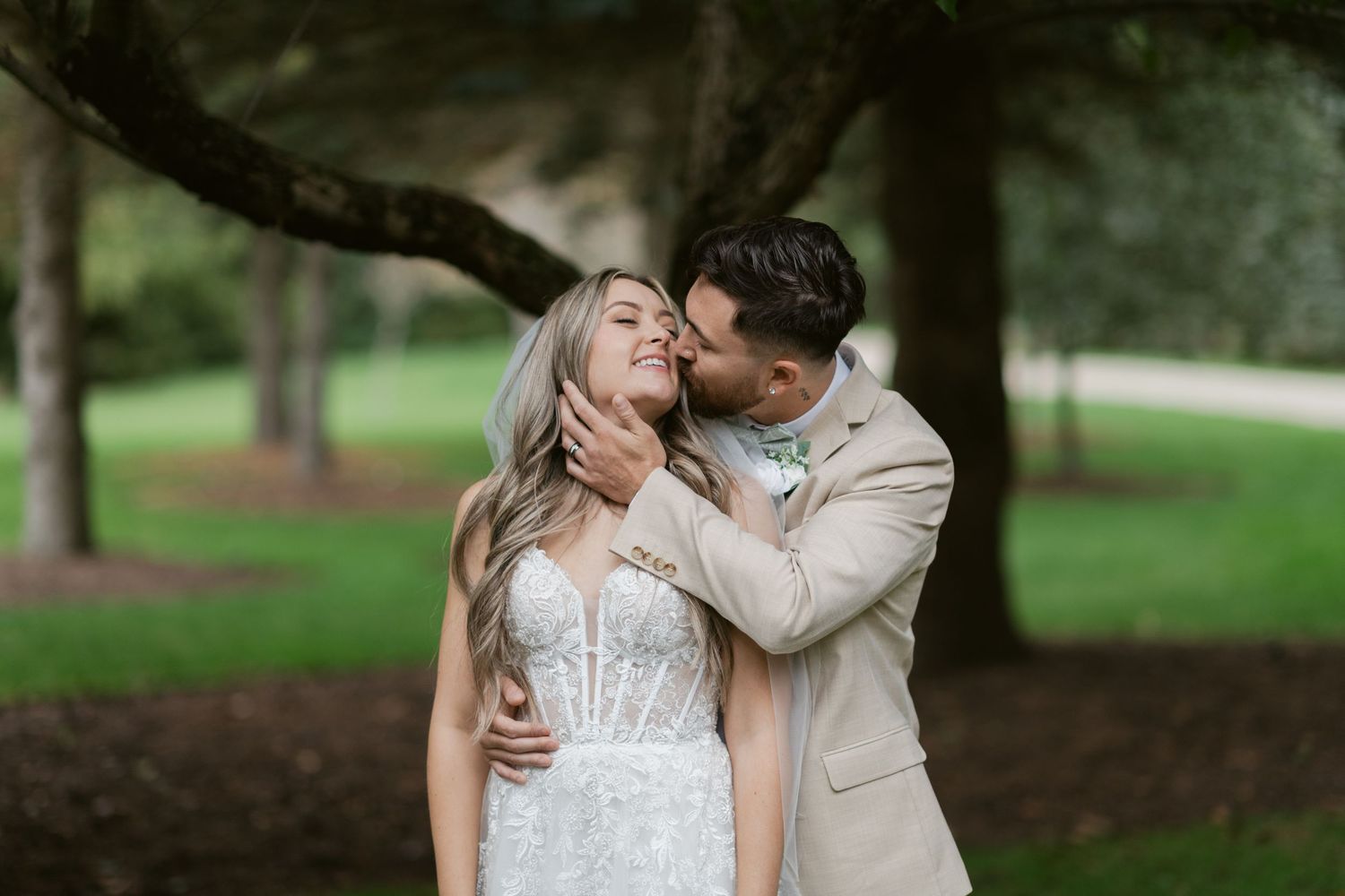 Groom in beige suit kissing bride in lace wedding dress under trees in outdoor garden setting.