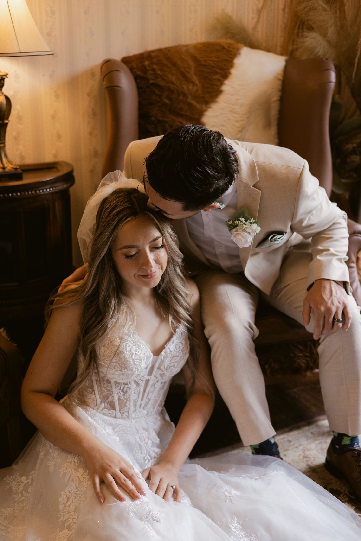 Groom in beige suit leans to kiss bride in lace gown as she sits on a vintage chair indoors.
