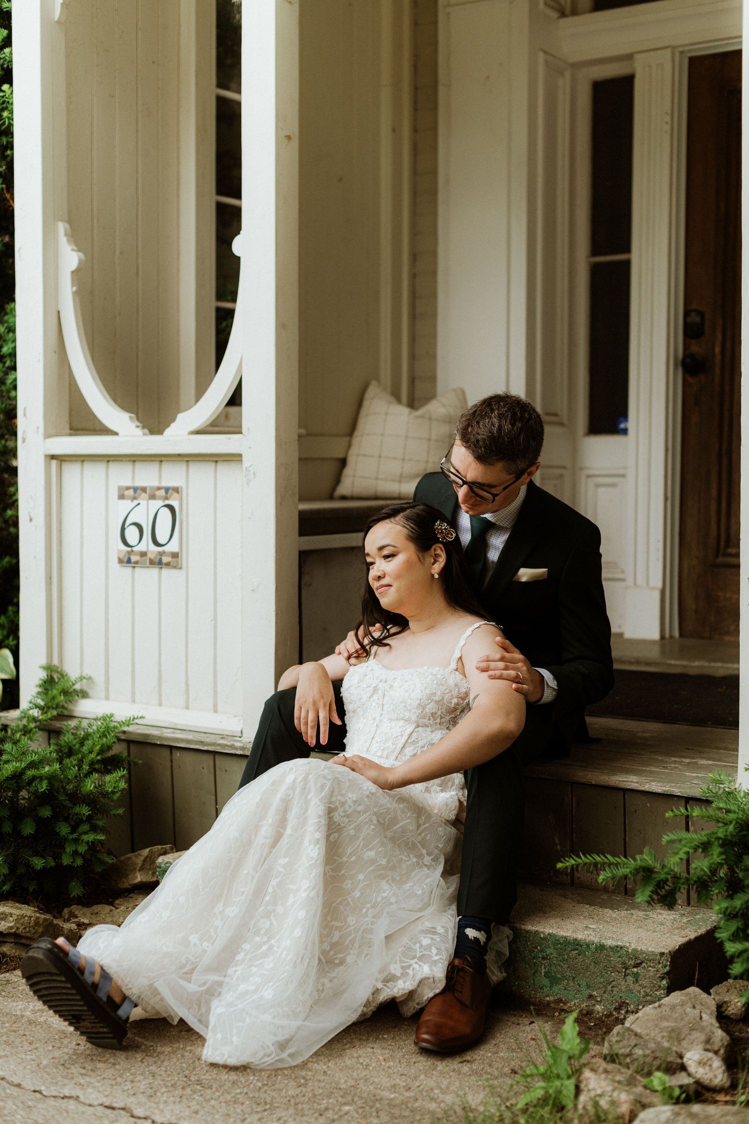 Bride in lace gown sits on porch steps with groom in black suit embracing her at house number 60.