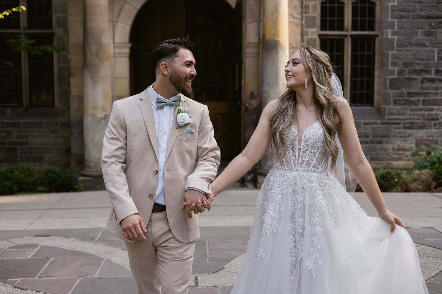 Bride and groom hold hands smiling in a sunlit stone courtyard, she in lace gown, he in beige suit with bow tie.