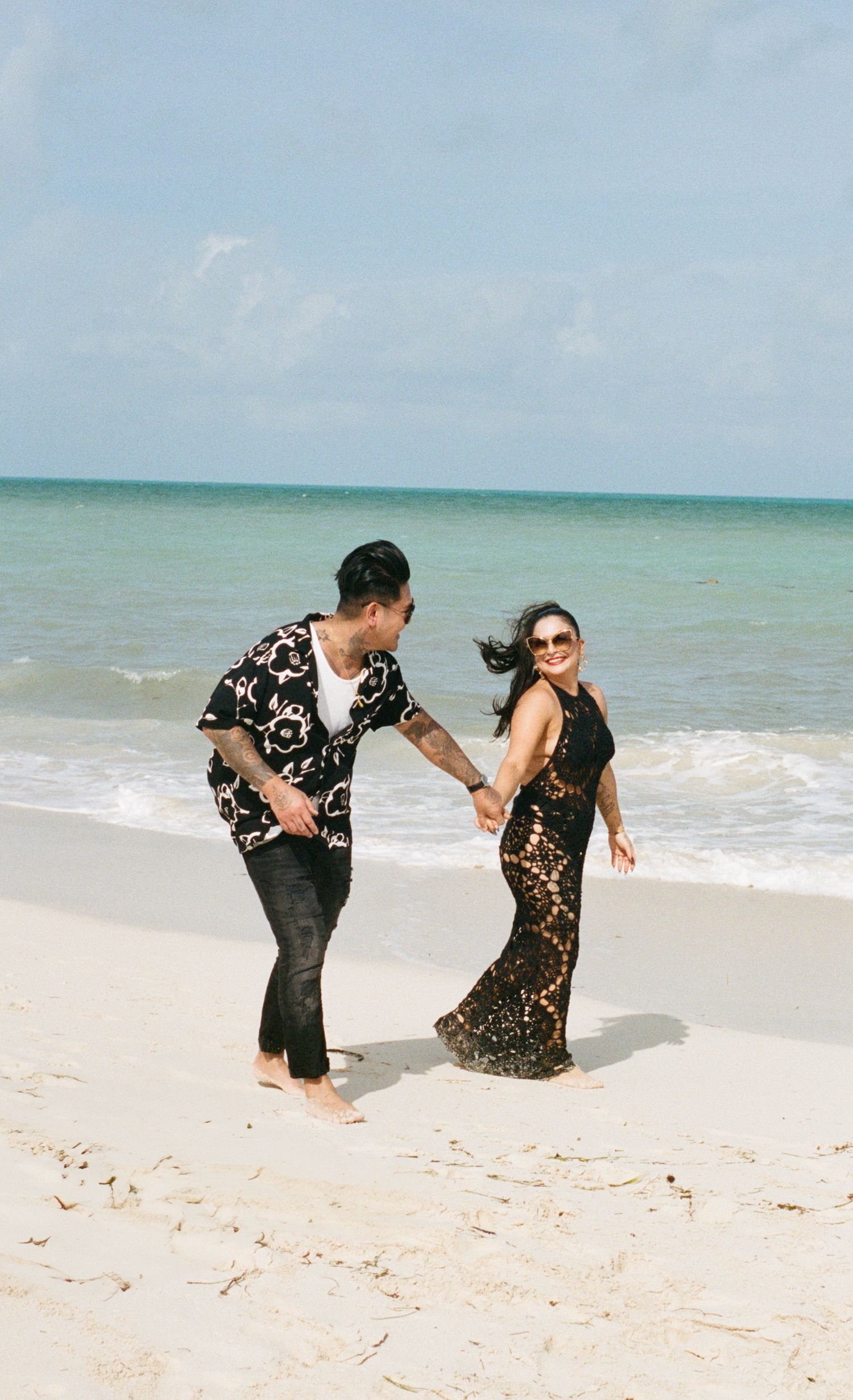 Couple dancing joyfully on a white sand beach with turquoise ocean waves, woman in black lace dress and man in floral shirt.
