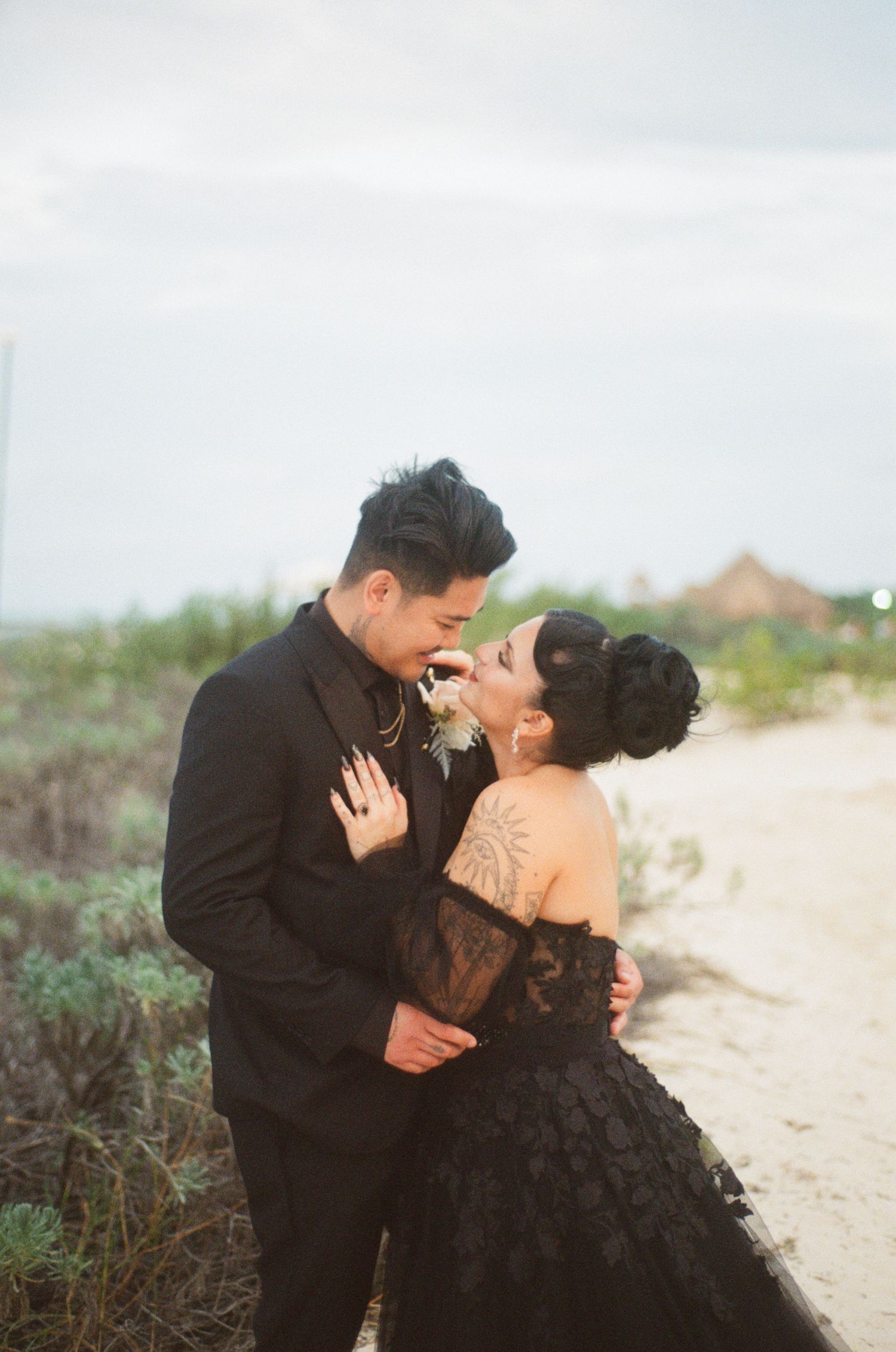 Laughing bride in black lace strapless gown with shoulder tattoo, groom in black suit, beach setting.