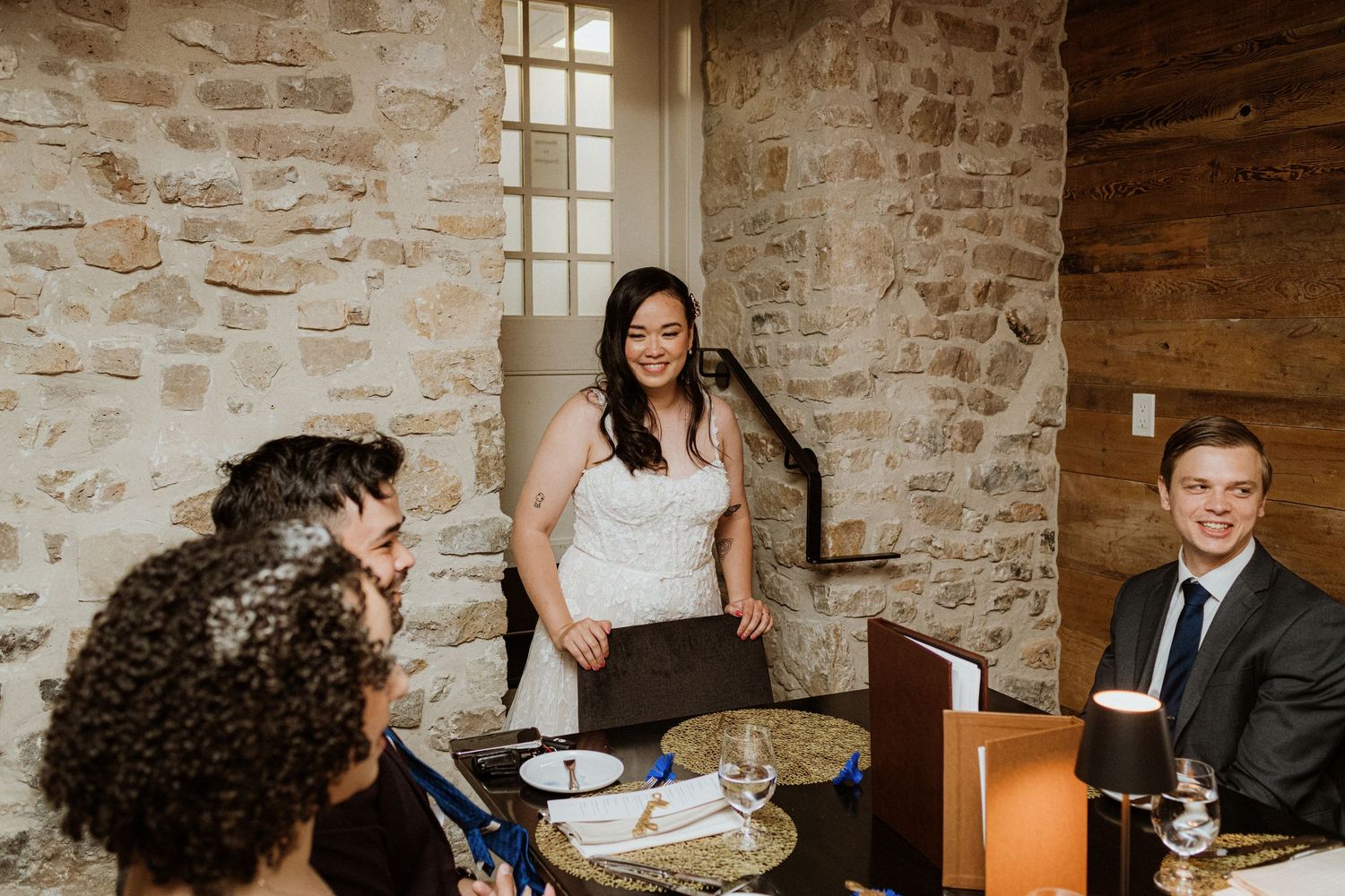 Bride in white lace dress standing and speaking to guests at an intimate wedding reception dinner.