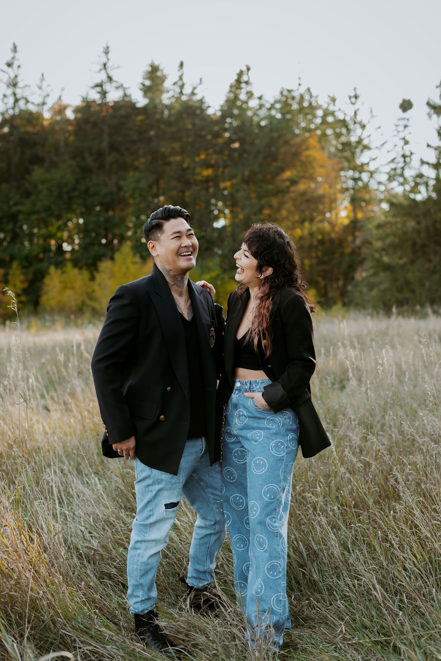 A happy couple in black blazers and jeans laughing together while walking through a meadow.