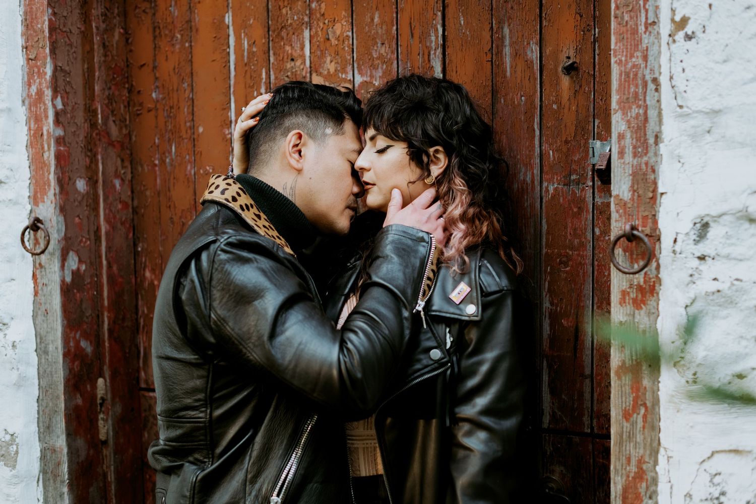 Couple in leather jackets sharing a romantic moment near a rustic wooden door.