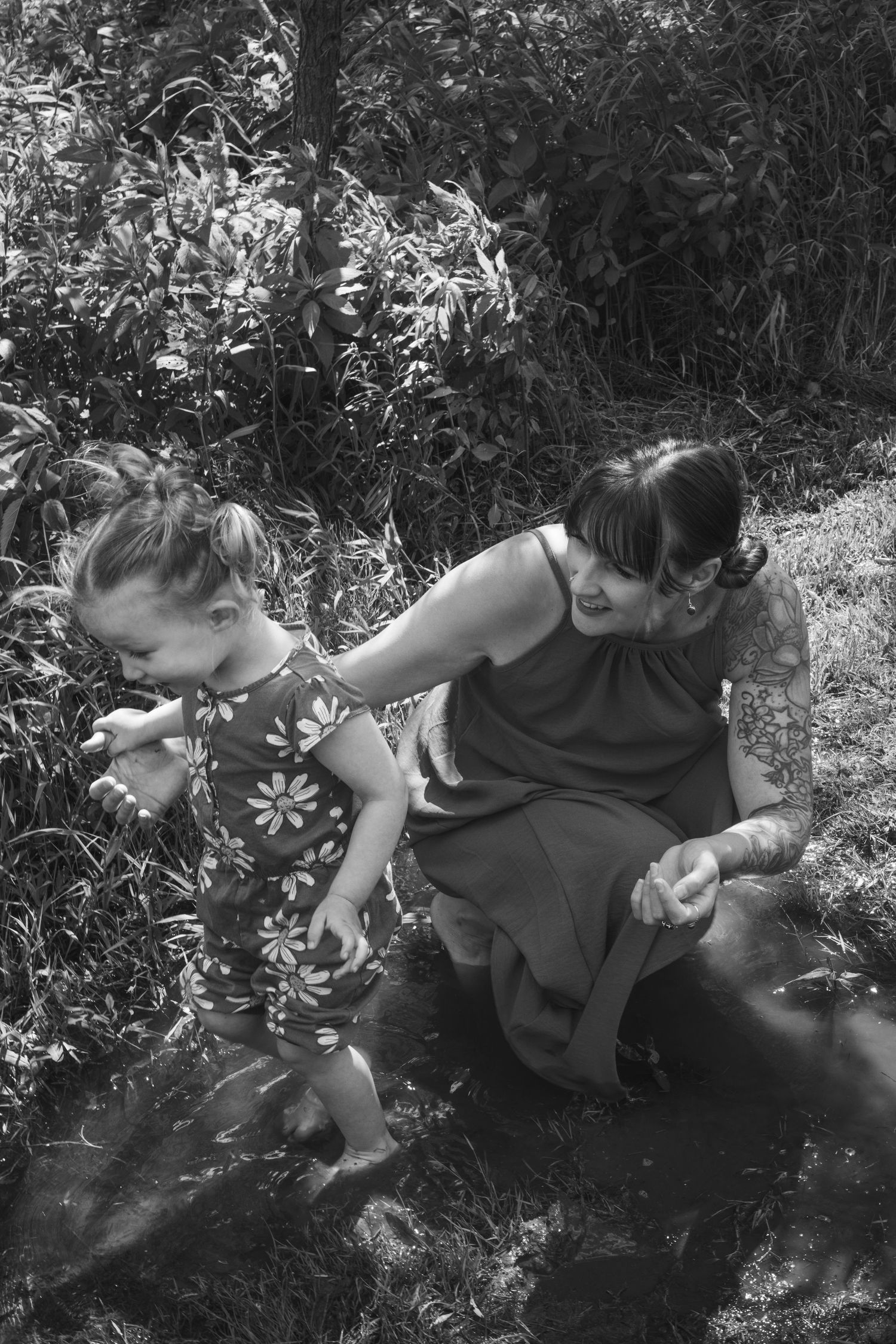 Black and white image of a tattooed mother sitting beside her toddler daughter playing near water in a garden.