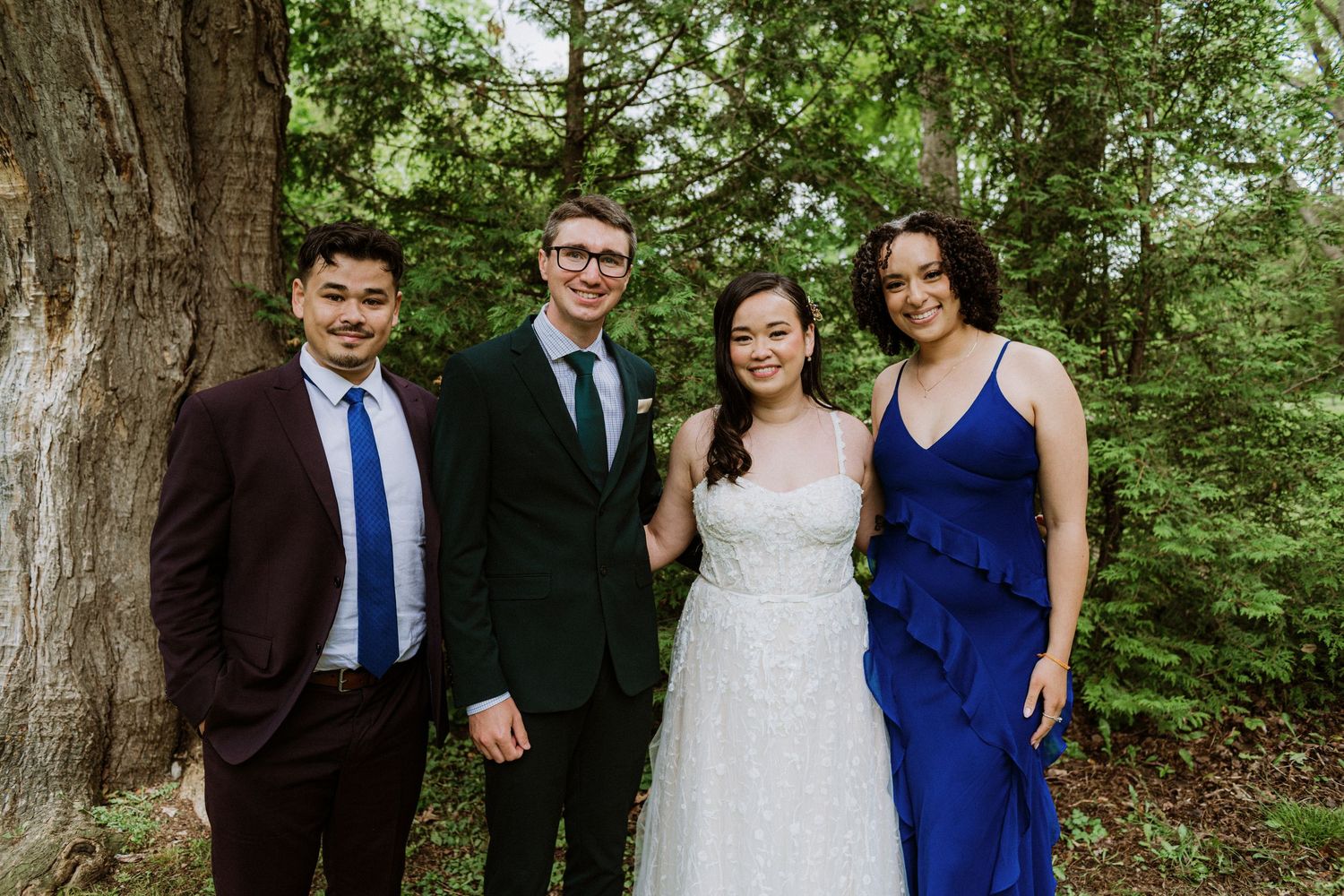 Bride and groom posing with two guests in blue and burgundy attire at outdoor wedding.