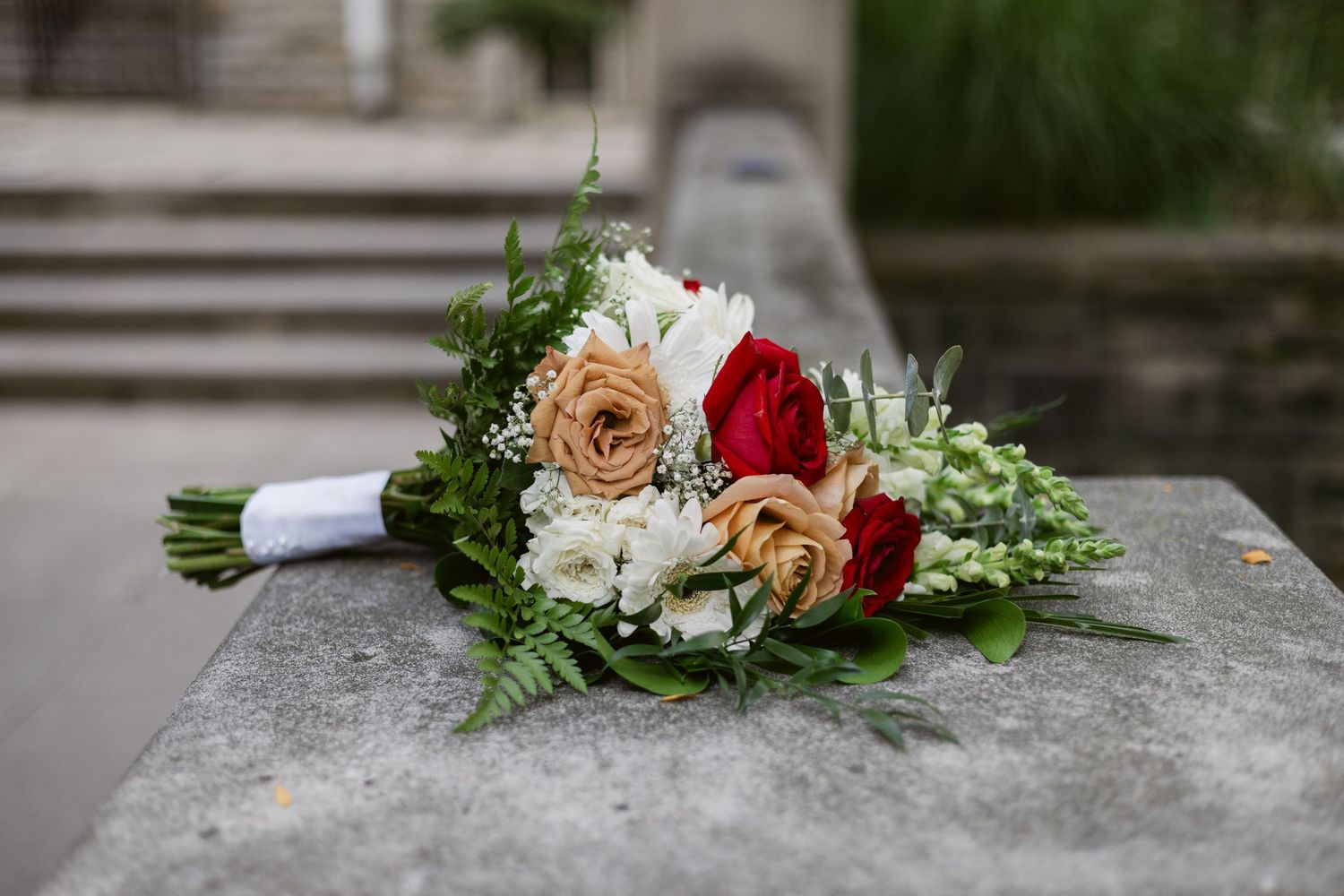Wedding bouquet of red, peach, and white roses with lush greenery resting on stone surface outdoors.