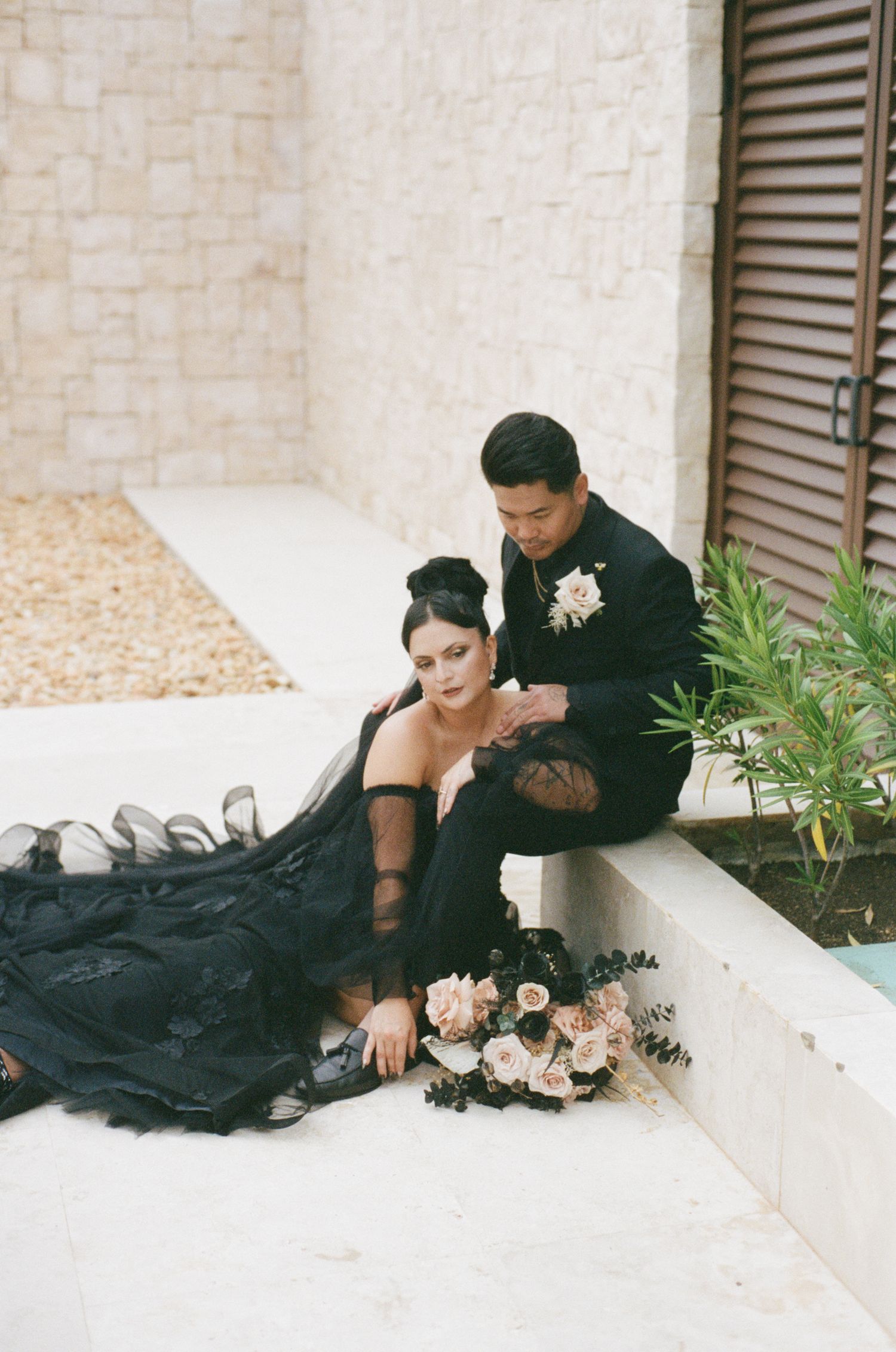 Wedding couple in matching black attire sit together on steps with blush and black bridal bouquet.