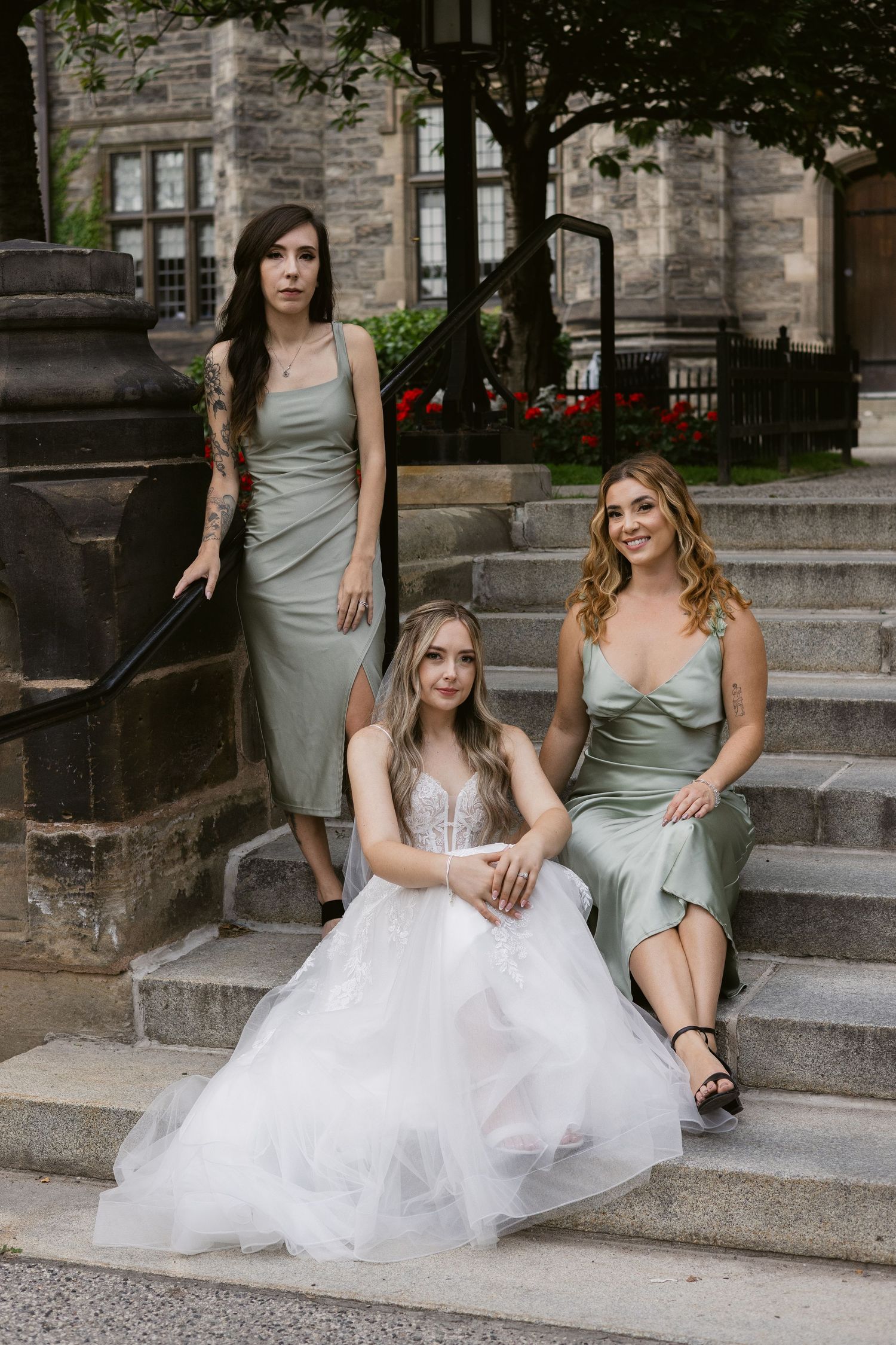 Bride in white tulle gown sits on steps with two bridesmaids in sage green dresses at stone building.
