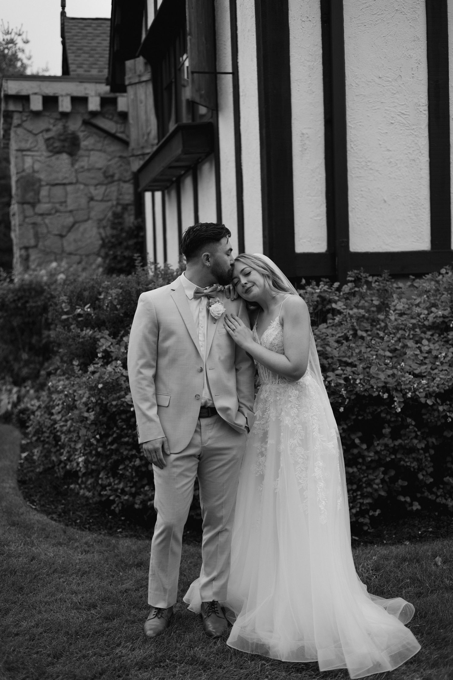 Black and white photo of groom kissing bride near Tudor-style building, bride in lace wedding gown.