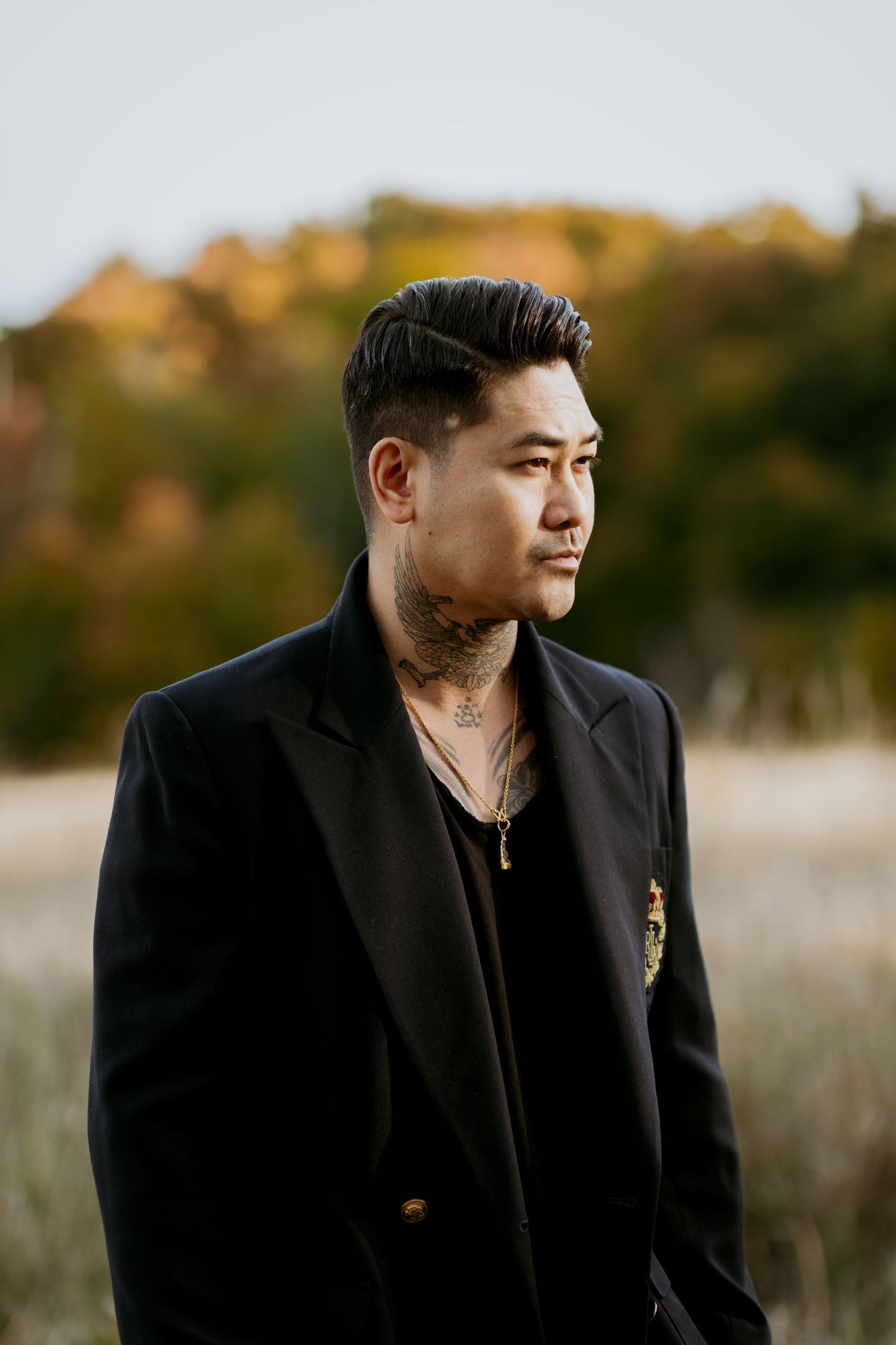 Stylish man with neck tattoos wearing black blazer and gold chain necklace posing outdoors in autumn setting.