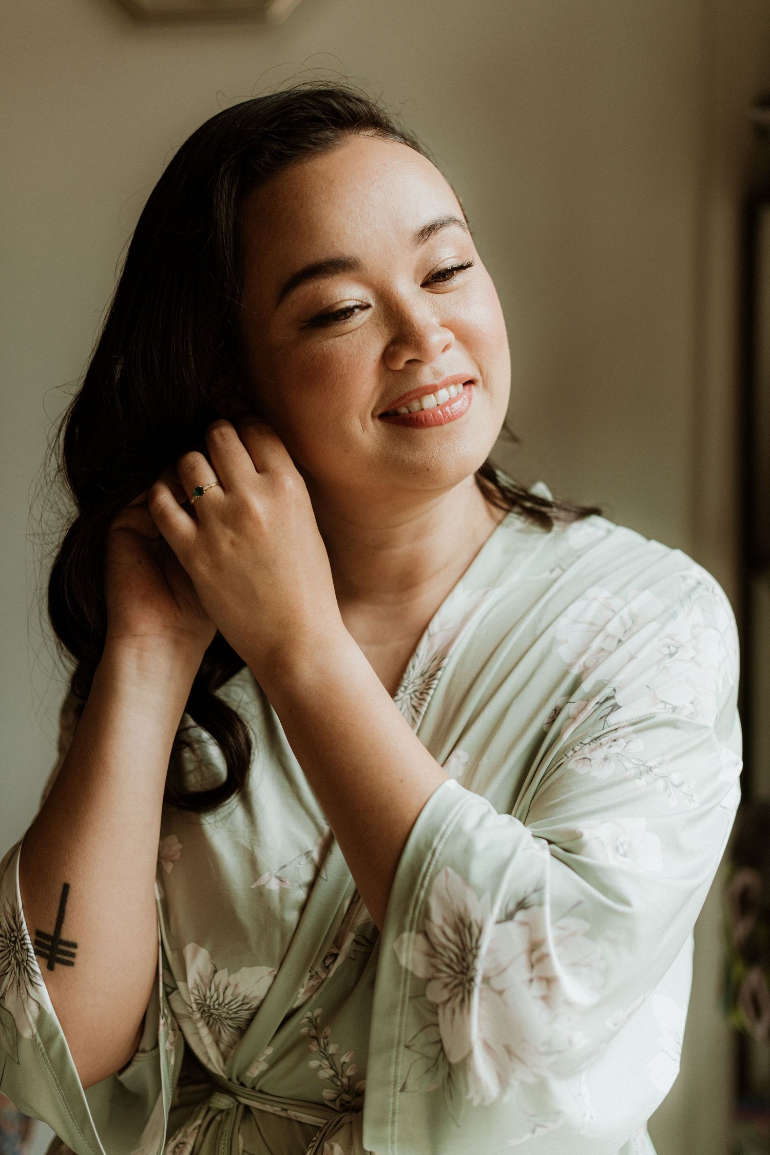 Smiling bride in sage floral robe touching her hair during wedding morning prep.
