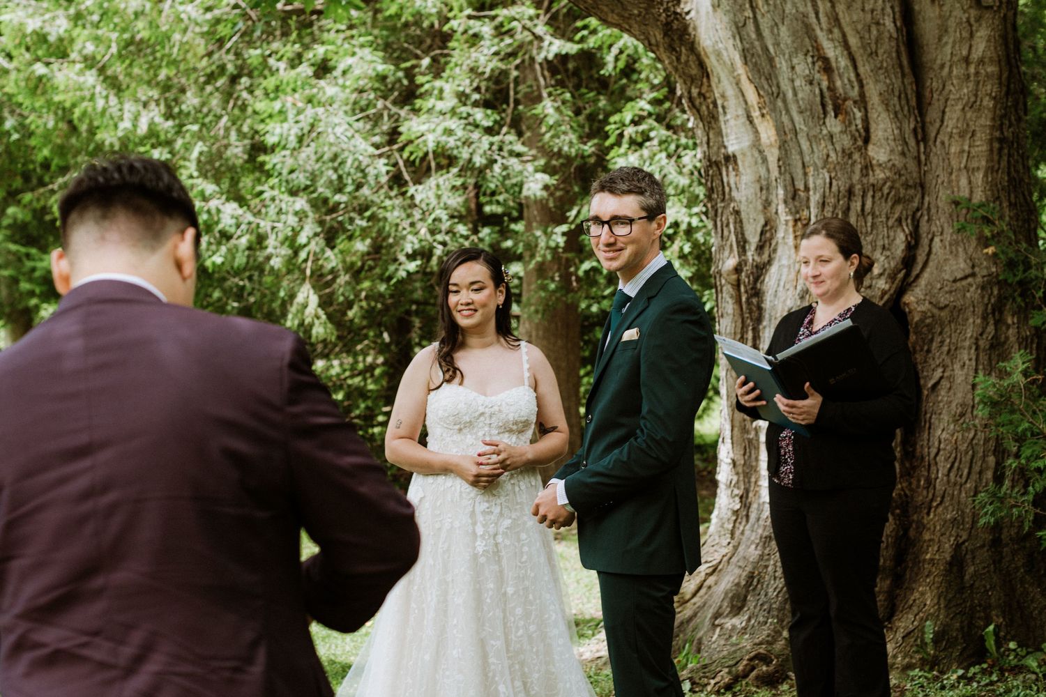 Guest reads during outdoor wedding ceremony as bride and groom stand with officiant near large tree.