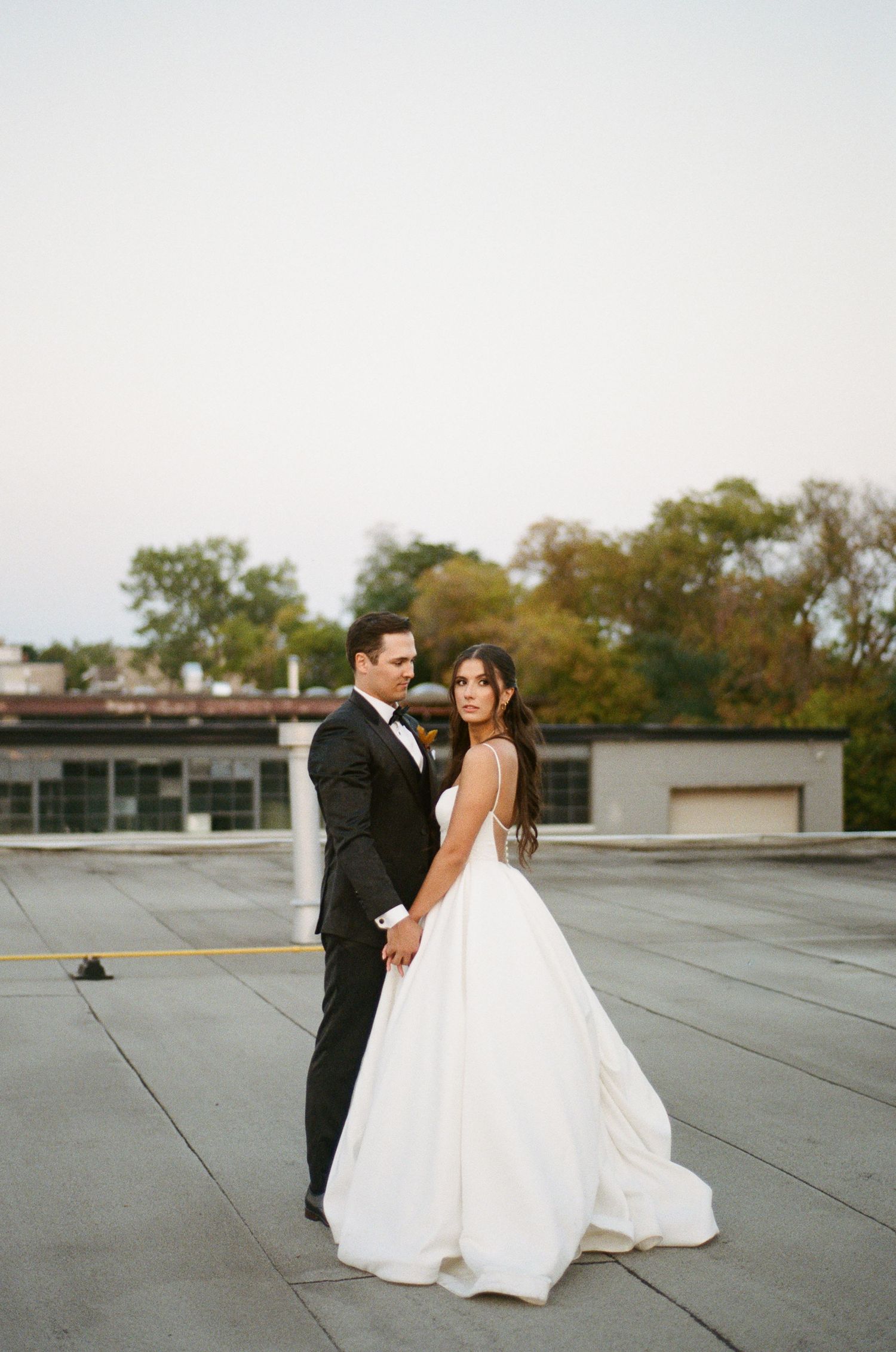 Elegant bride in white ball gown poses with groom on rooftop, looking back at camera.