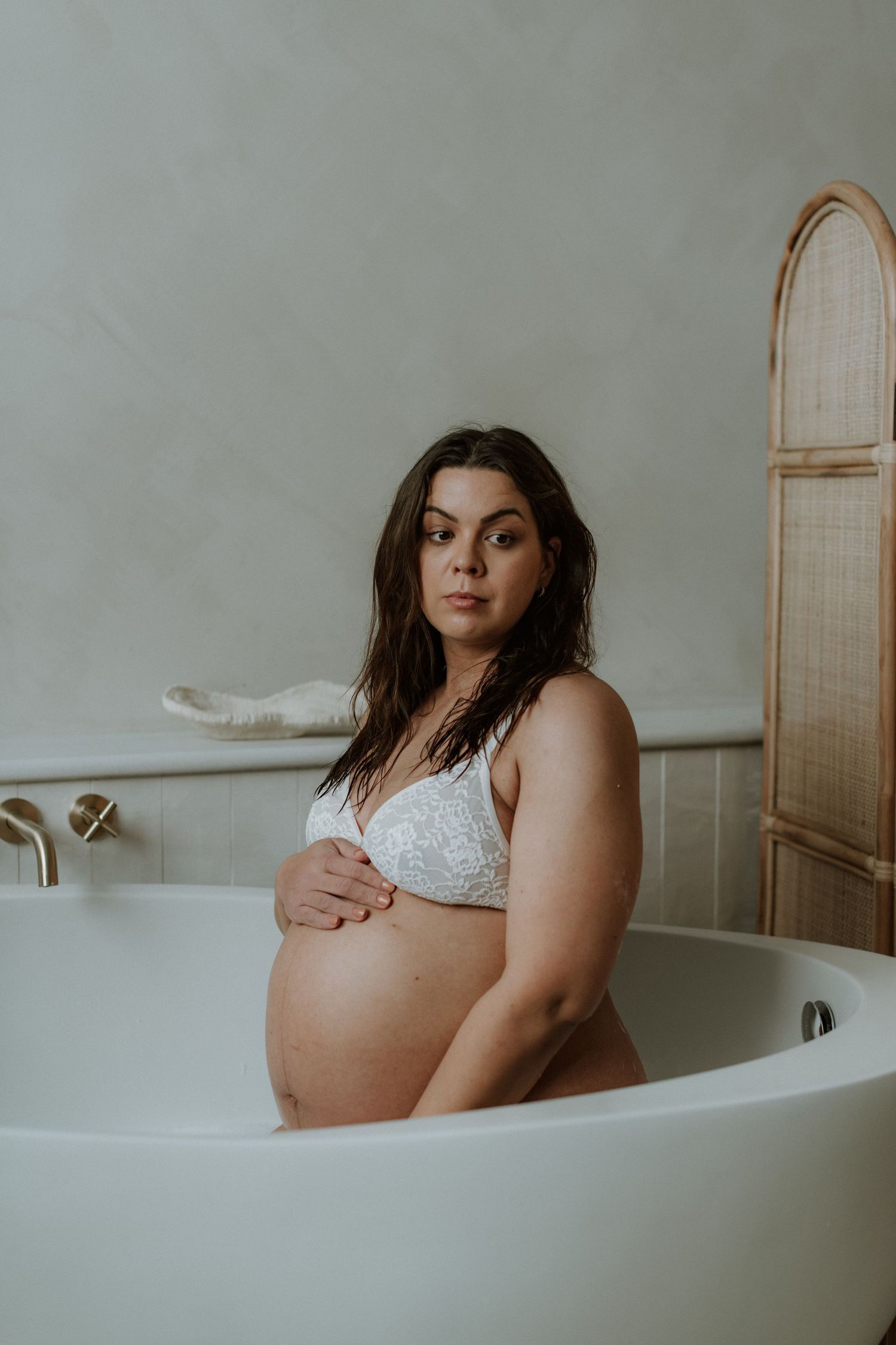 Pregnant woman in white lace bra sitting in a freestanding bathtub, cradling her baby bump.