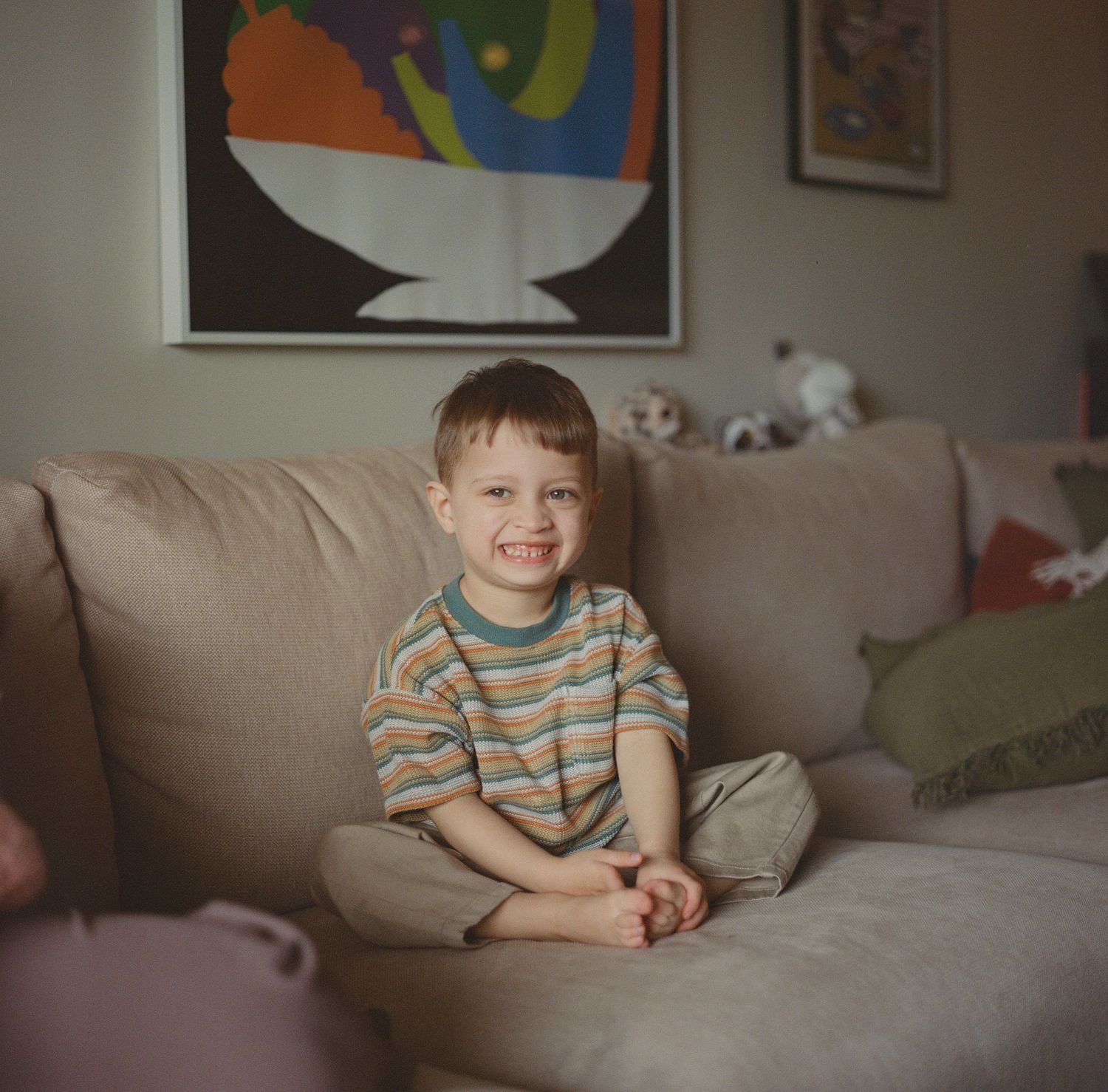 Smiling young boy in striped shirt sits cross-legged on sofa with colorful artwork behind him.