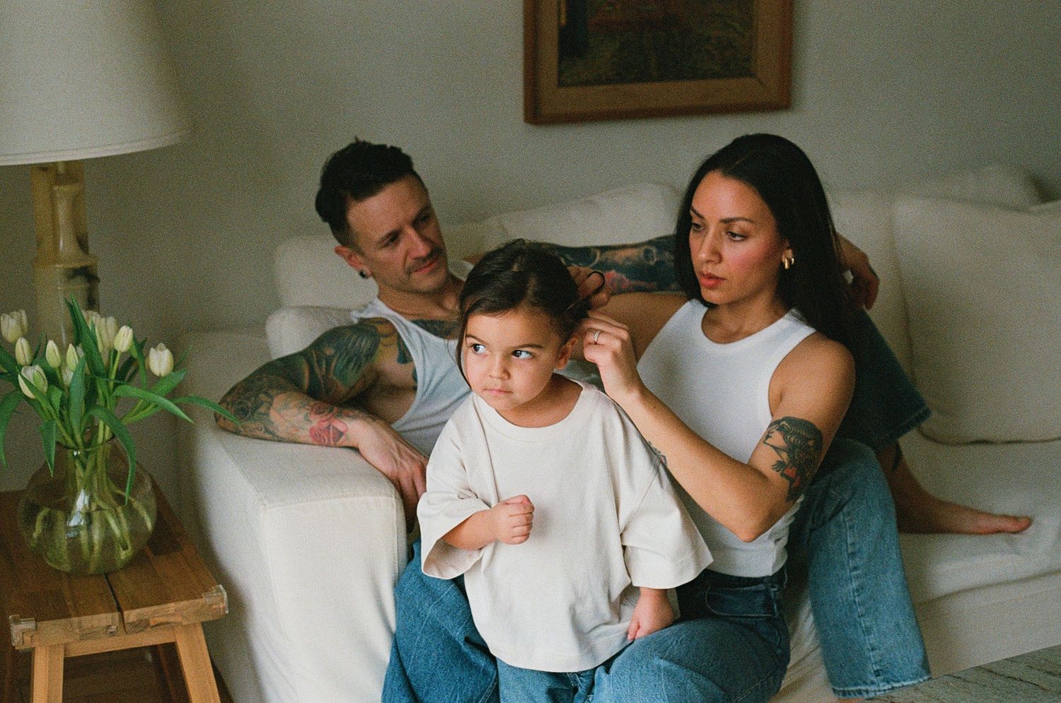 Tattooed mother styling young daughters hair while father smiles on white sofa at home.