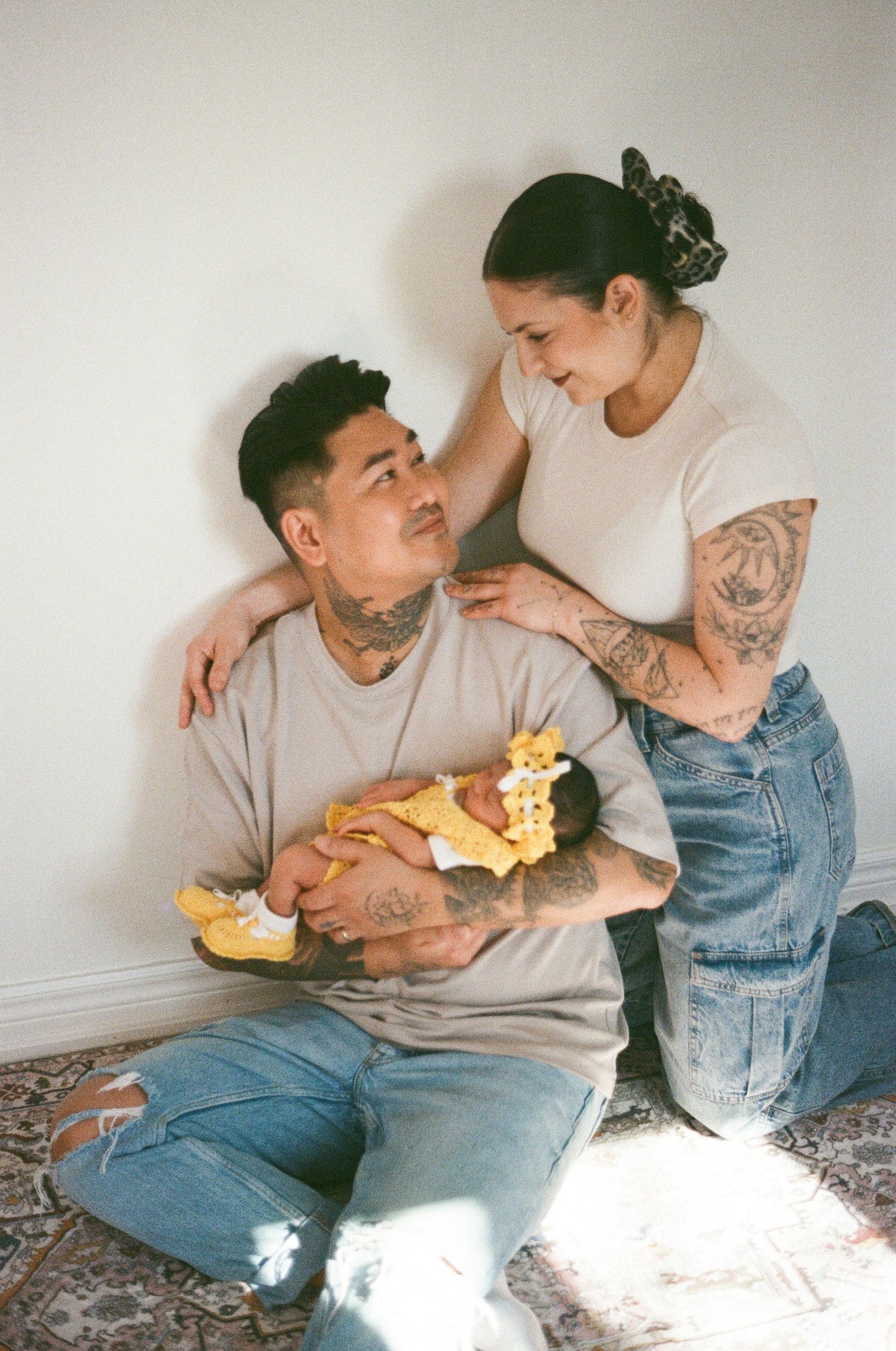 Tattooed parents sitting against a white wall, father cradling their newborn in yellow outfit.