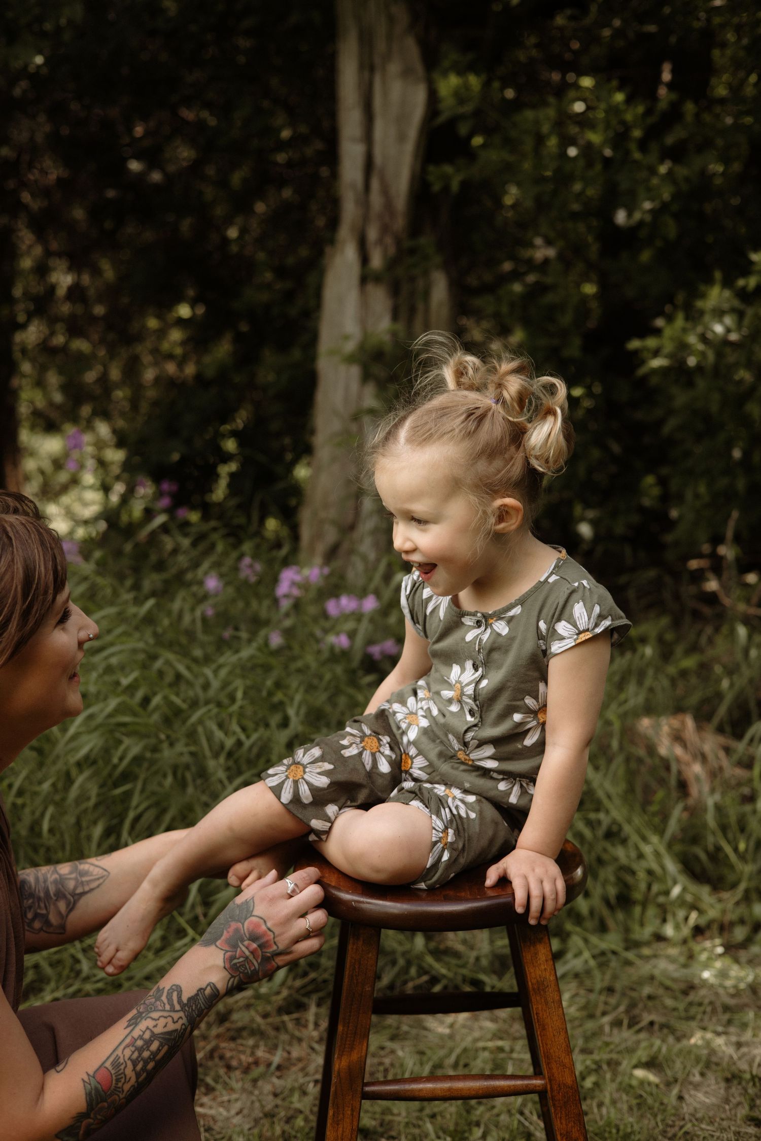 Smiling toddler girl in daisy print romper sits on wooden stool while tattooed mother kneels beside her outdoors.