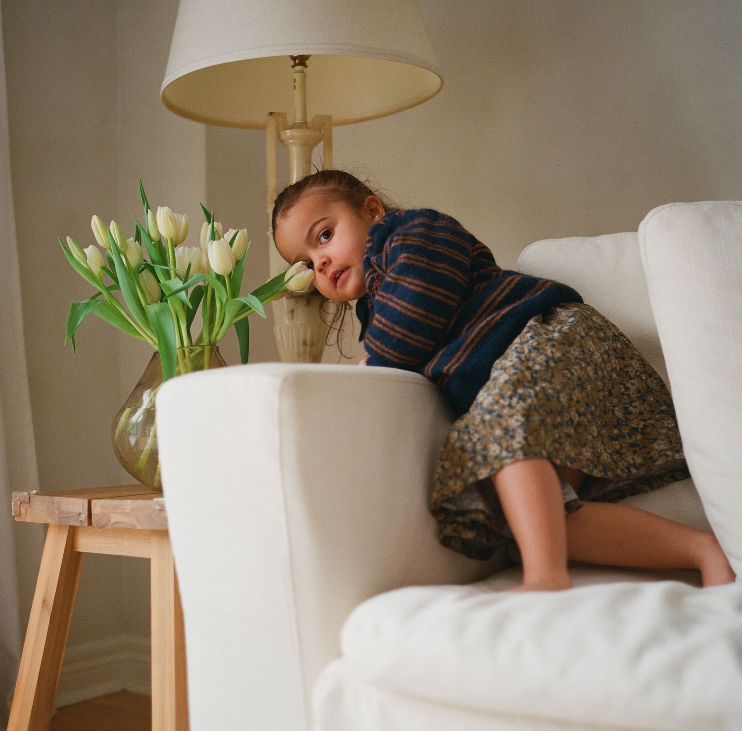 A young child drapes over the arm of a white armchair beside a vase of white tulips on a wooden stool.
