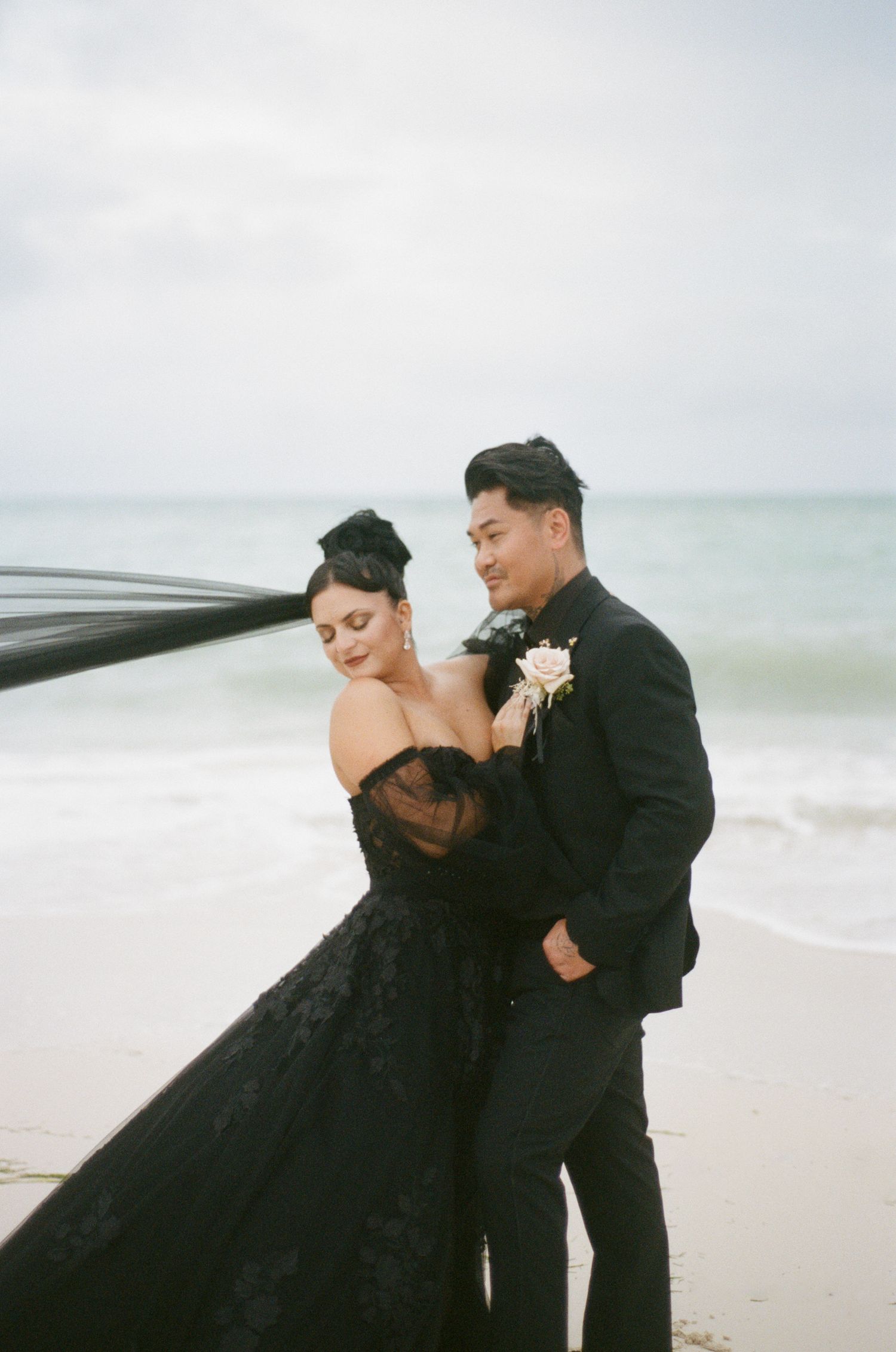 Couple embracing on beach, black veil flowing in wind, groom in black suit with boutonniere, ocean backdrop.