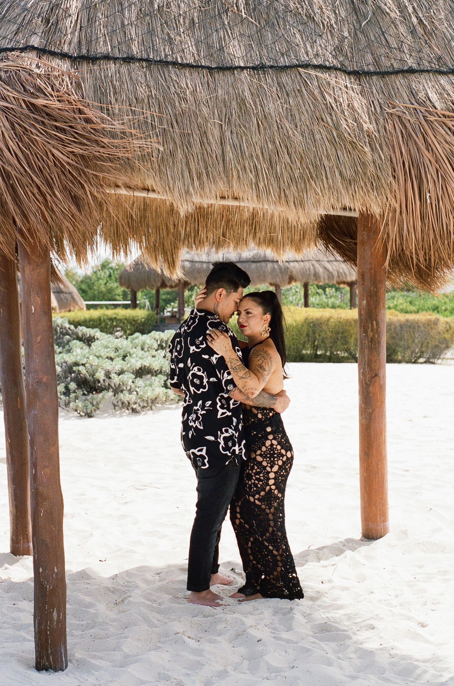 Romantic couple embracing under a thatched palapa on white sand beach, woman in black lace dress.