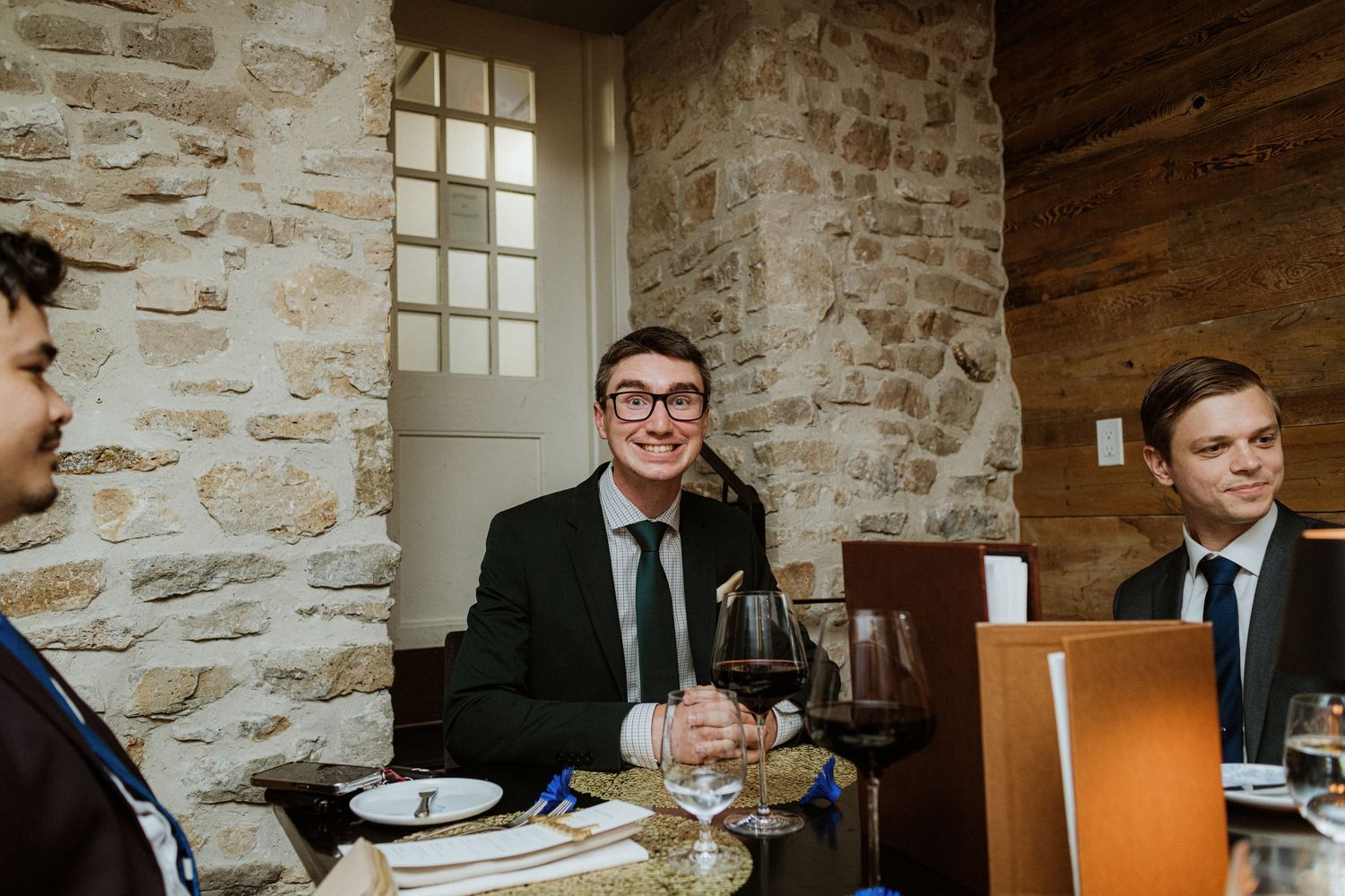 Young man in suit and glasses smiling at wedding reception dinner table with stone wall background.