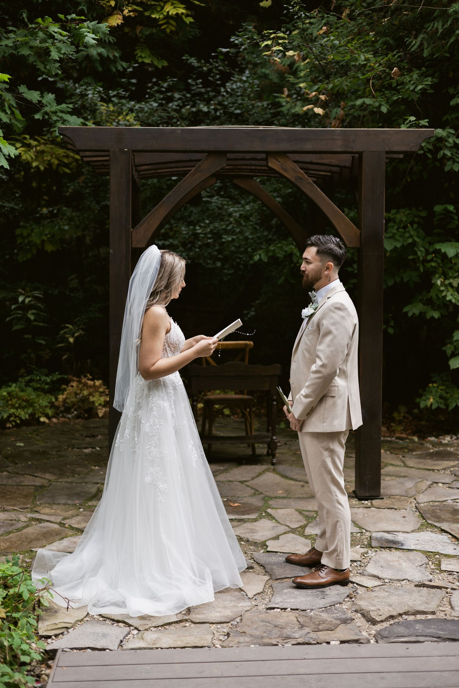 Bride reading vows to groom under rustic wooden arch on stone patio surrounded by lush greenery.