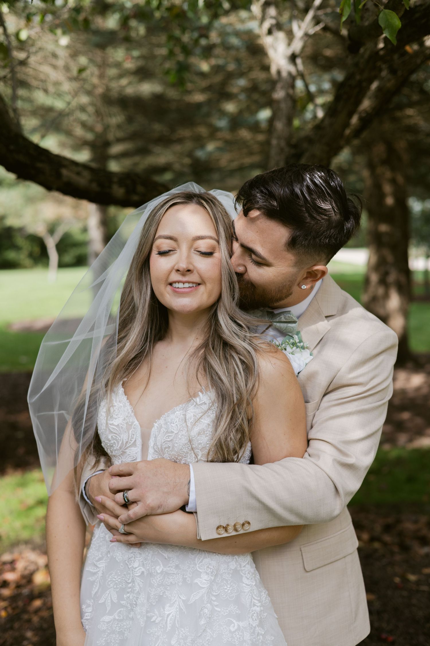 Groom kisses bride on cheek from behind under a sheer veil in a lush green garden setting.