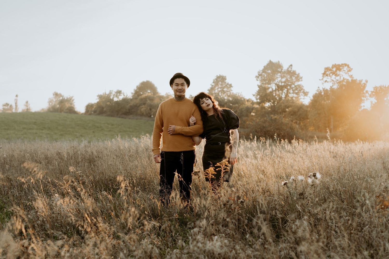 Couple standing together in glowing sunset field, man in mustard sweater and black hat beside woman in dark outfit.