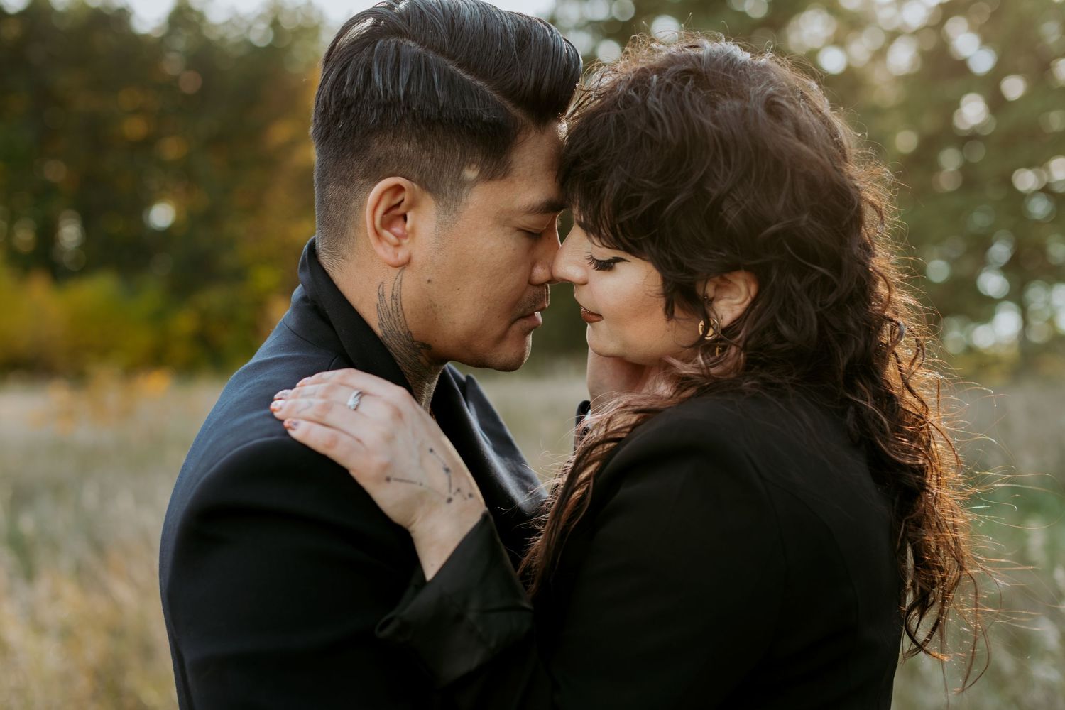 A romantic couple in black outfits sharing an intimate forehead touch in a golden field.