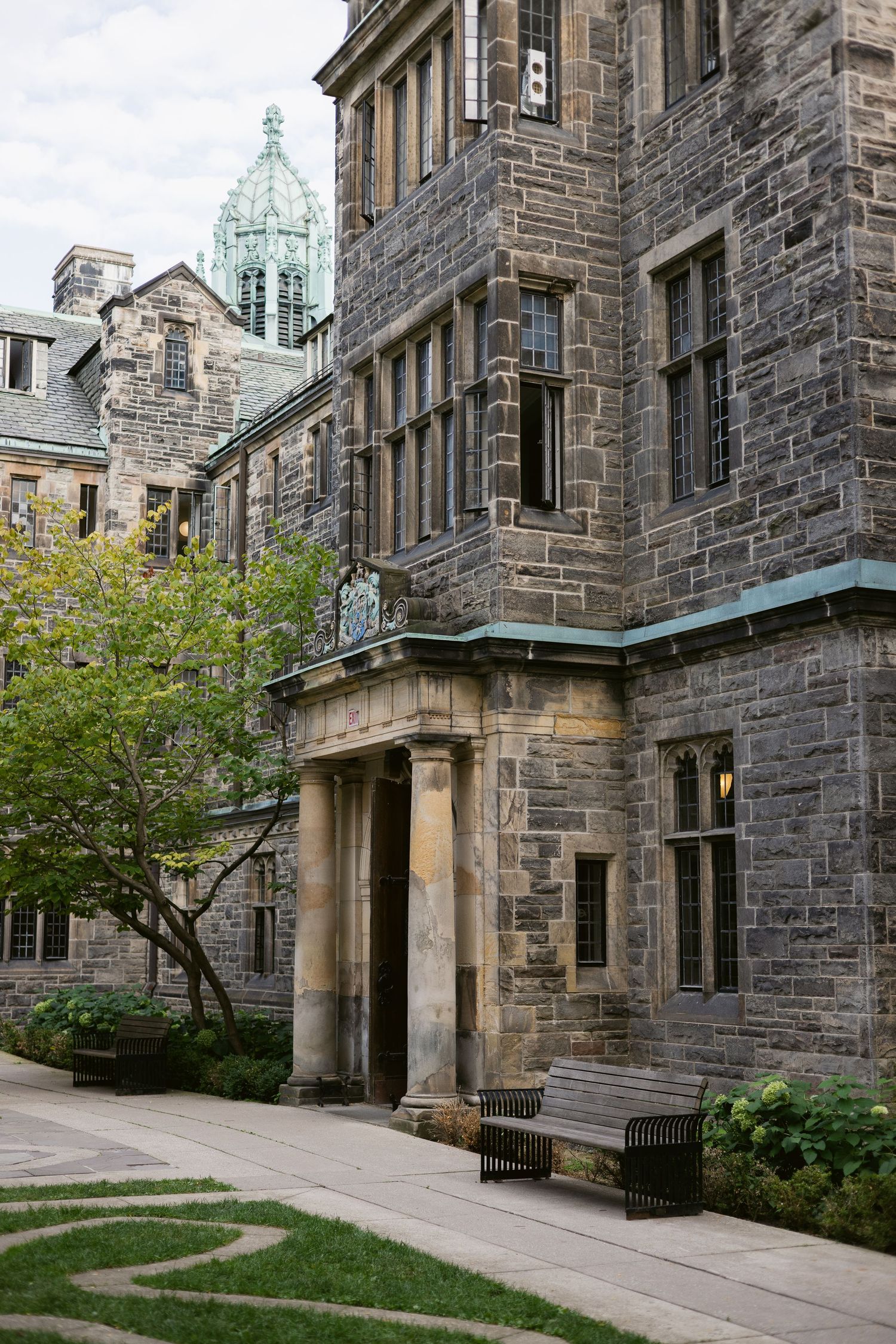 Corner entrance of a Gothic stone building with arched doorway, copper roof tower, and manicured garden pathway.