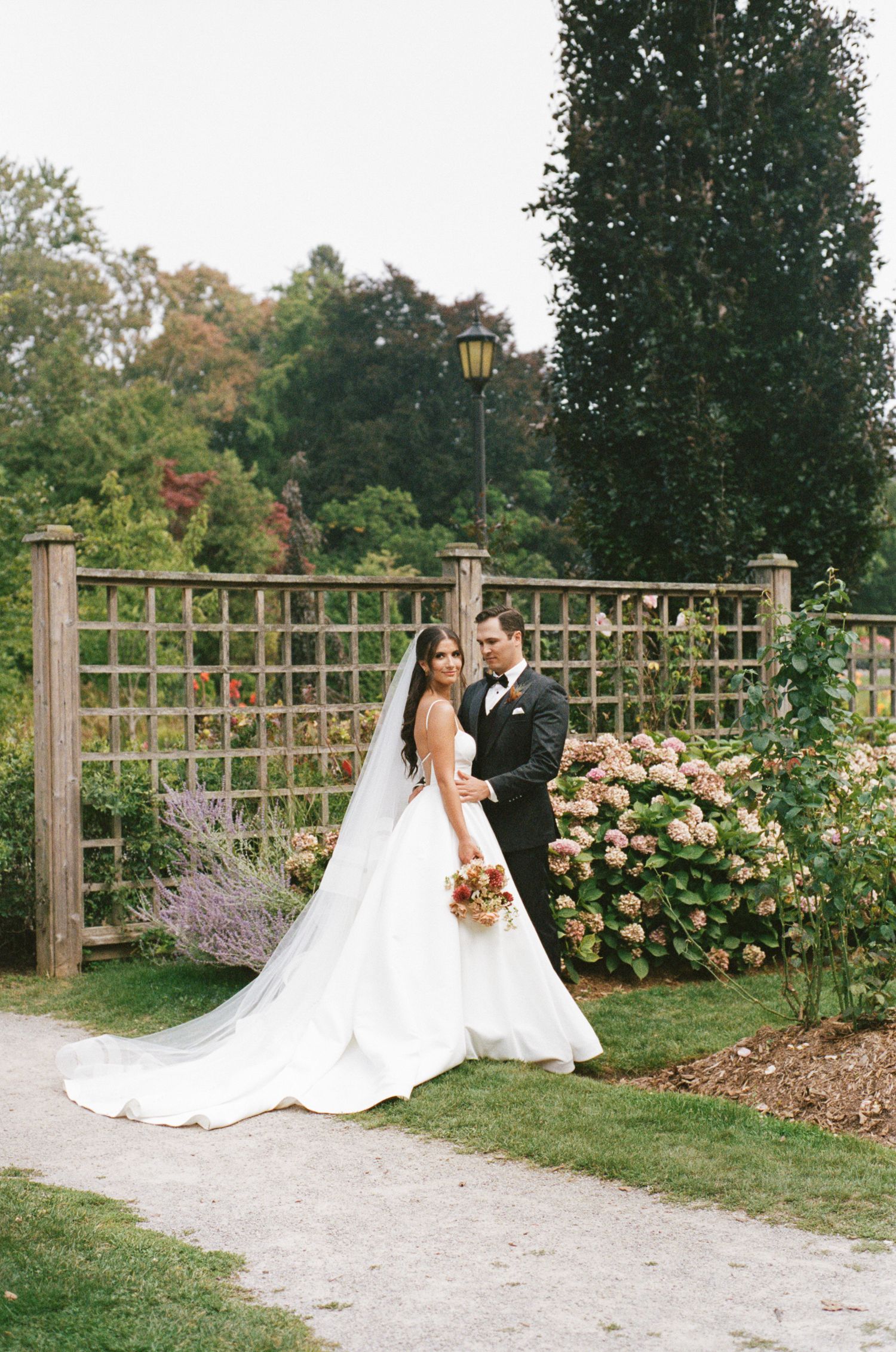 Bride and groom posing in a lush garden with hydrangeas and a wooden trellis fence on their wedding day.