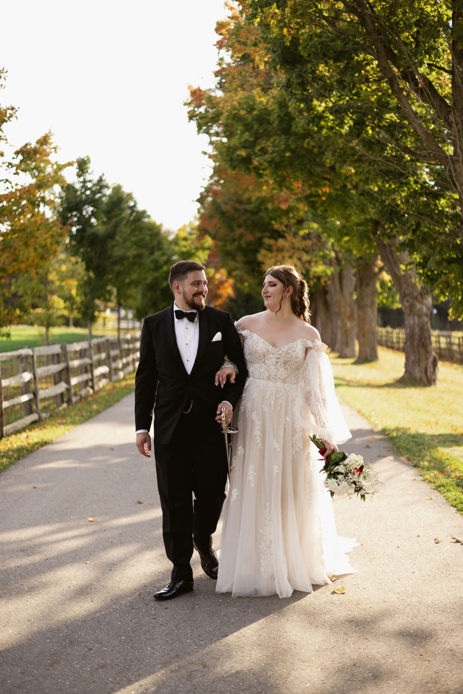 Bride and groom walking along a tree-lined path in autumn, bride holding a bouquet.