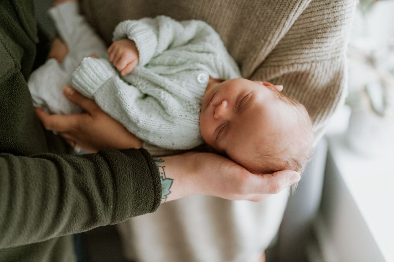 Sleeping newborn baby in a knit sweater cradled gently in the arms of a parent wearing a green sweater.